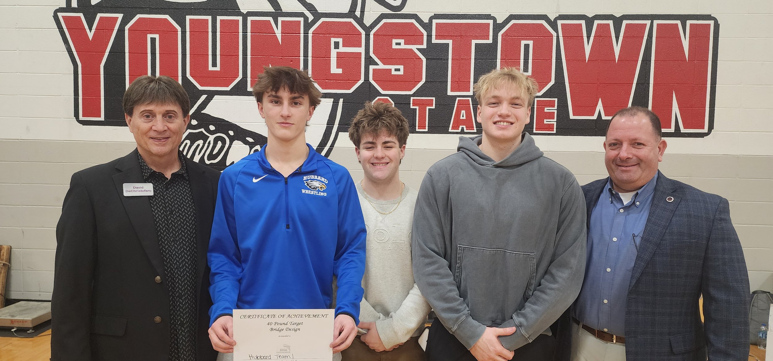 Group of five people standing in front of a wall with a big sign banner reading 'Youngstown' and 'Wrestling'. The two adults on each end are dressed in suits, and the three young men in the middle are holding a certificate of achievement and smiling.