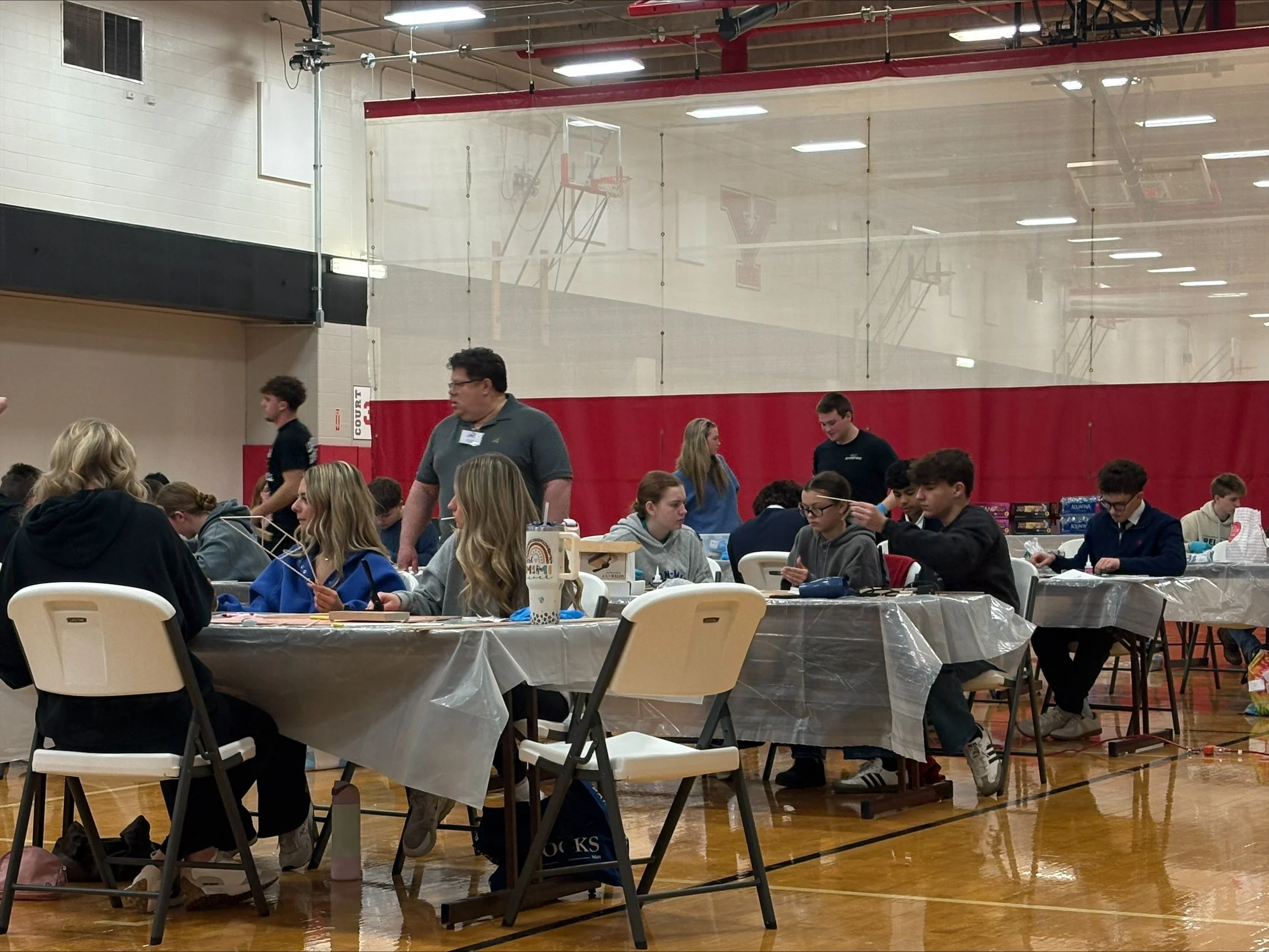 A gymnasium filled with people sitting at folding tables covered with plastic tablecloths, engaging in activities. In the background, basketball hoops are visible behind a clear divider. The ceiling has red beams and fluorescent lighting. Some indivi
