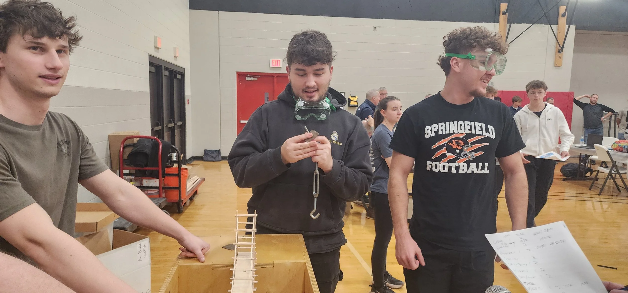 Group of young people gathered in a gymnasium, participating in a bridge-building competition, with participants working on their projects and observing others.