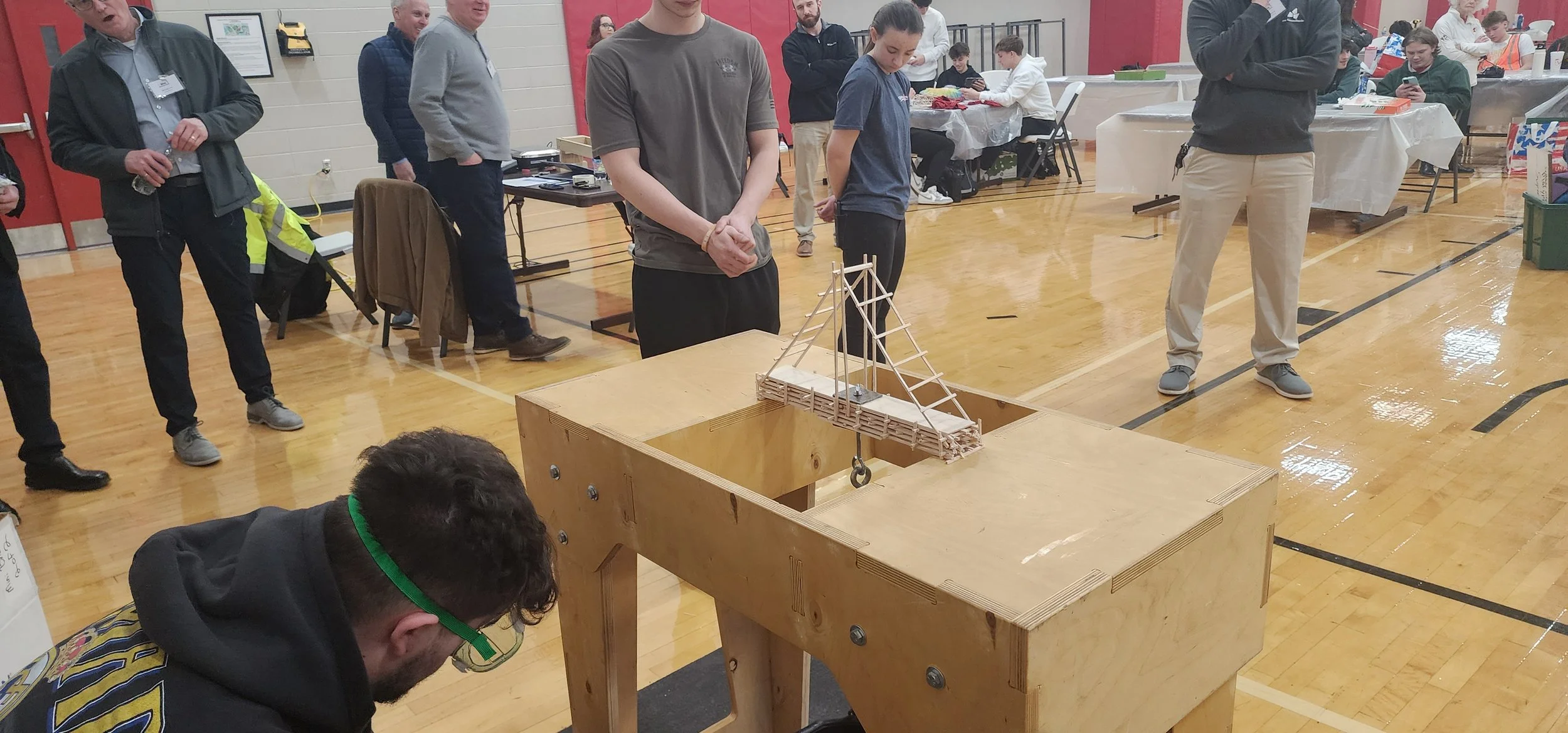 People gathered in a gymnasium watching a small wooden bridge model being tested with a pulley system.
