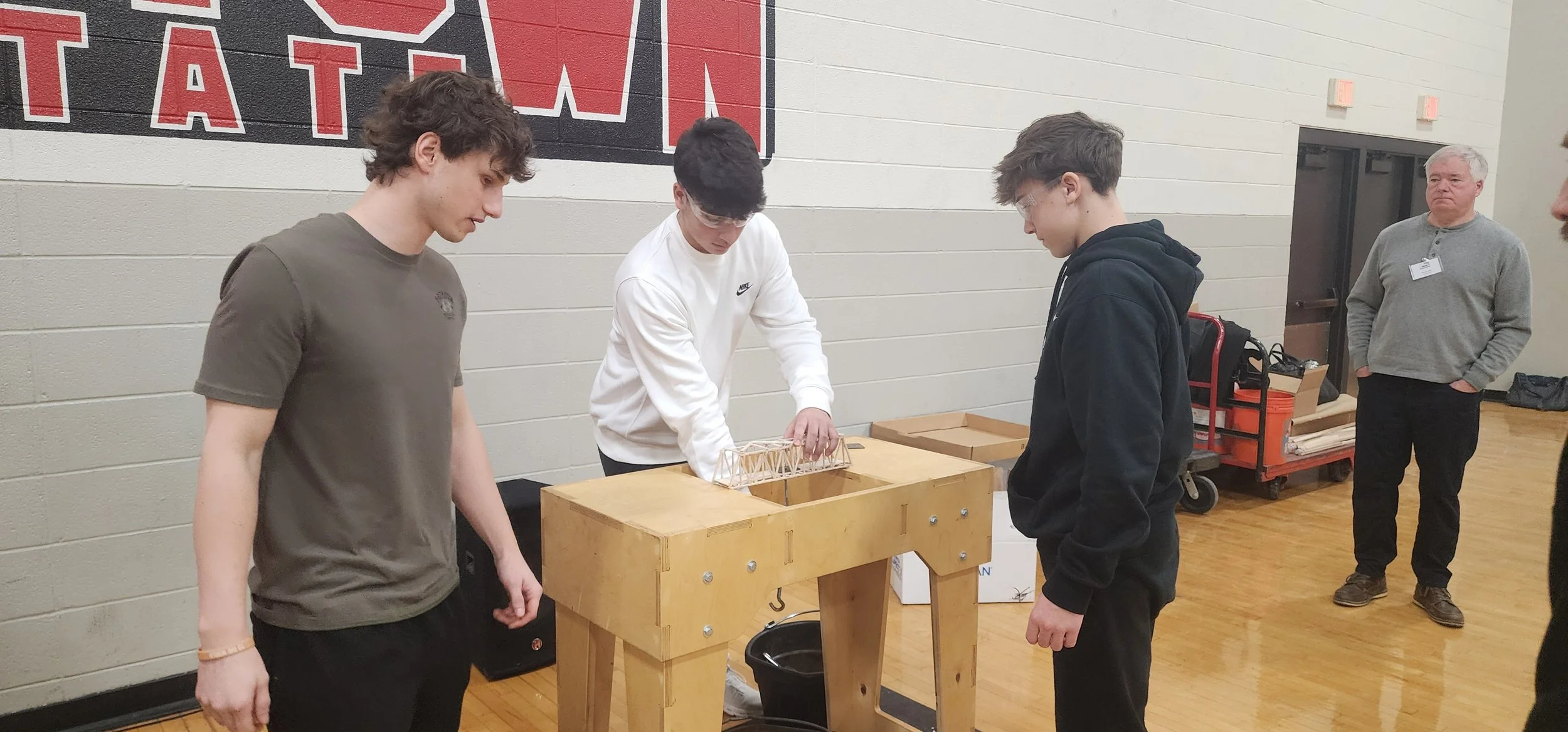 Three young men are inspecting a small model of a bridge on a wooden work table, with a person observing and another adult standing in the background in a gymnasium or community center setting.