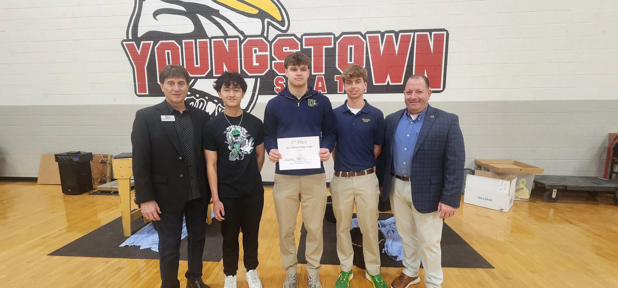 Group of five people standing inside a gymnasium in front of a large wall logo of Youngstown State University, with two students in the center holding an award certificate for second place in a bridge design contest.