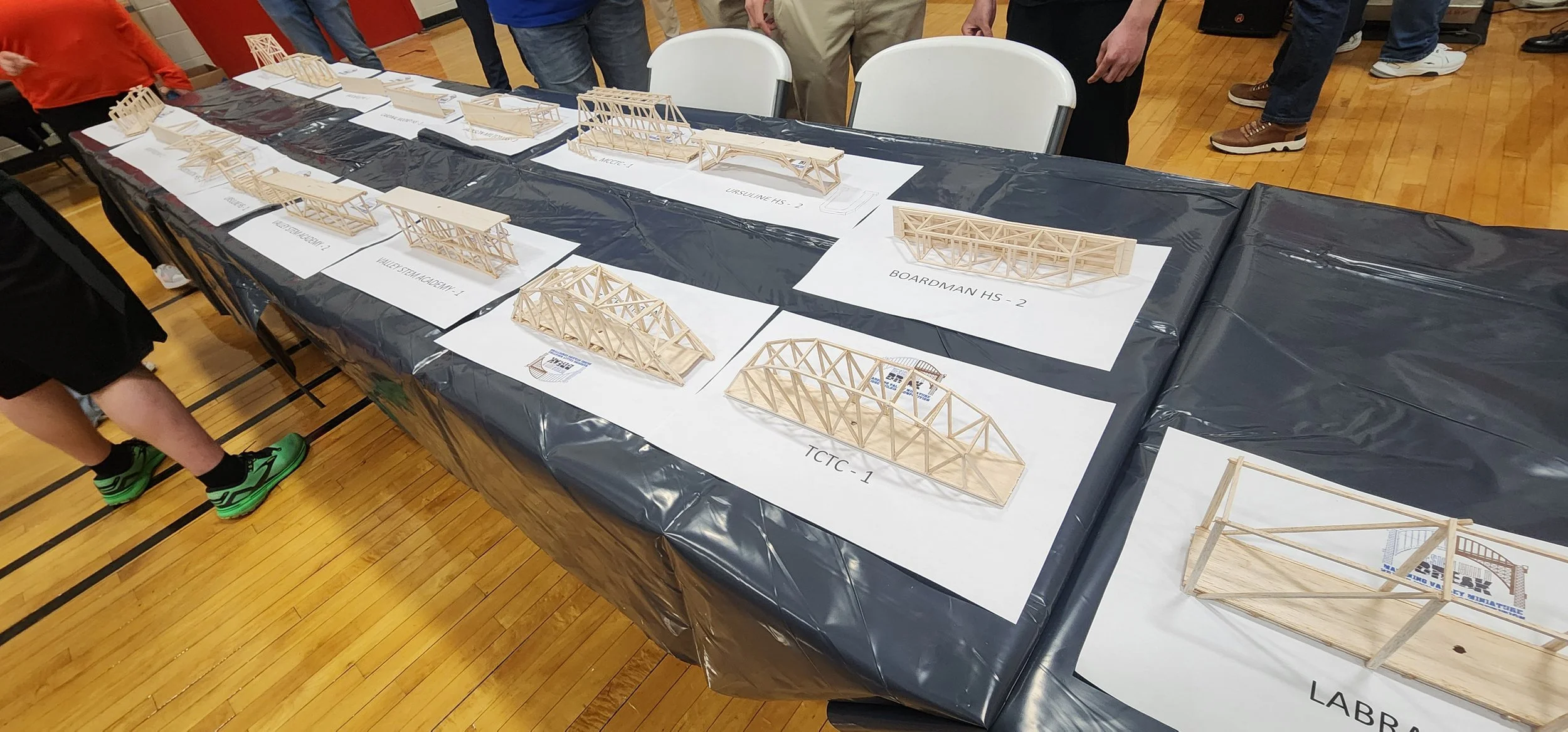 Table with multiple architectural model structures made of wooden sticks, labeled with project names, in a gymnasium with a wooden floor and people standing around.