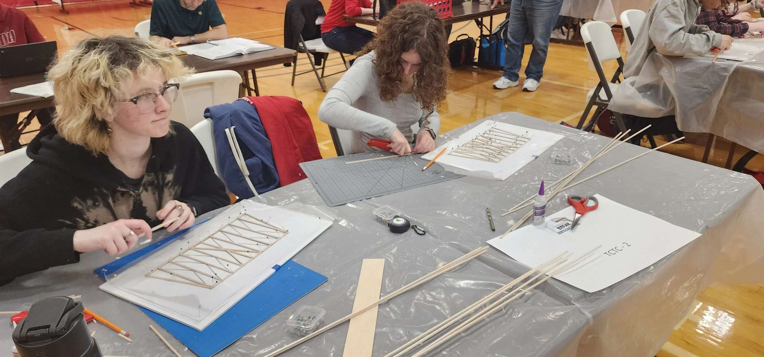 People participating in a workshop or class at a table with various craft supplies, including wooden sticks, glue, scissors, and tools, in a gymnasium or community center.