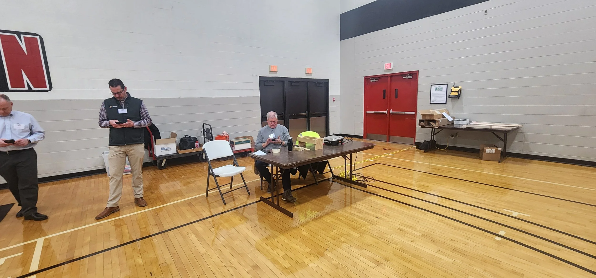 A gymnasium with a wooden floor, two men standing on the left using smartphones, a woman sitting at a table using a phone, various boxes and equipment on and near the table, opposite double red doors, and a table with more boxes on the right.