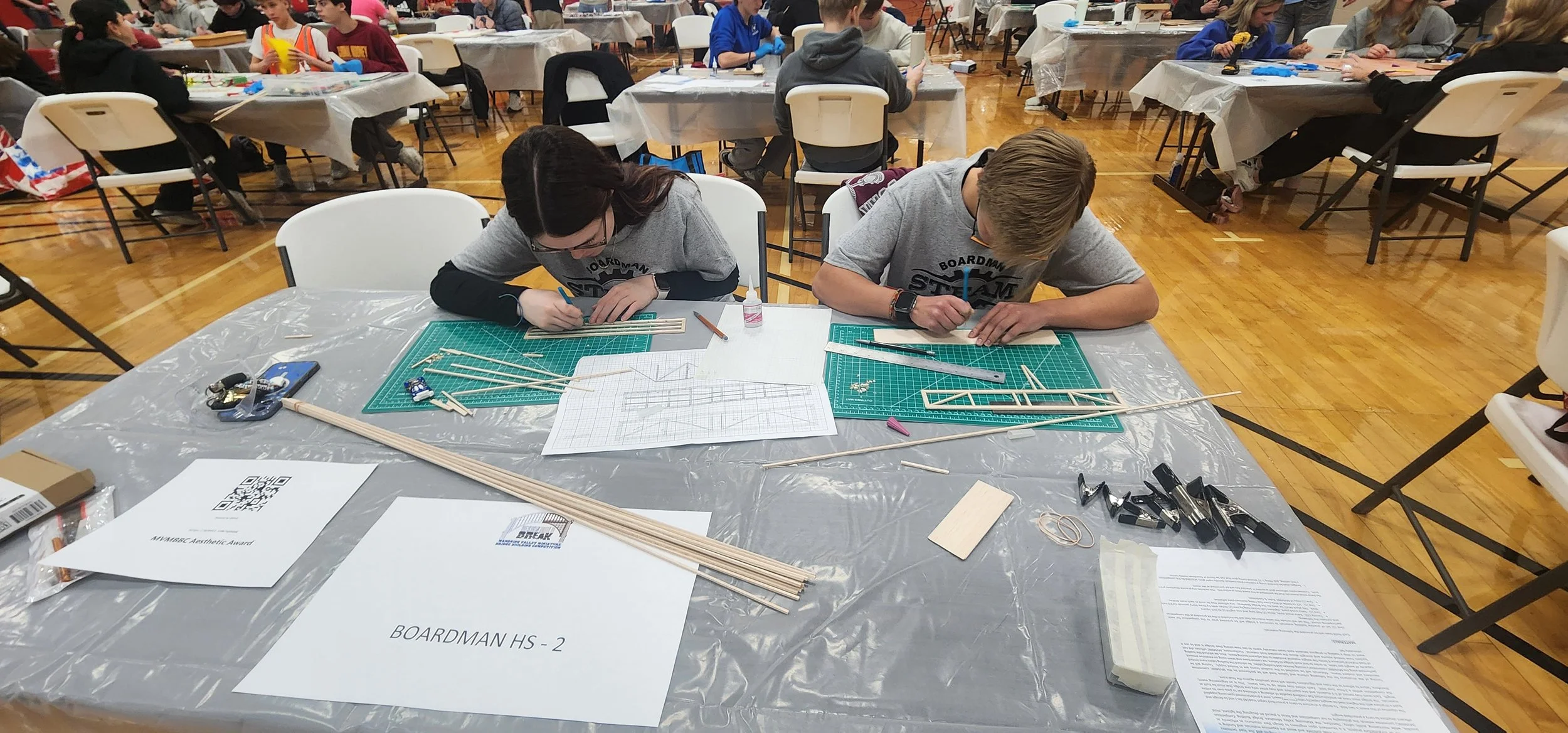 Two students, a girl with dark hair and a boy with blonde hair, are working on a model project at a table covered with tools, wooden sticks, paper, and drawing lines, in a large classroom or gymnasium with other students in the background.