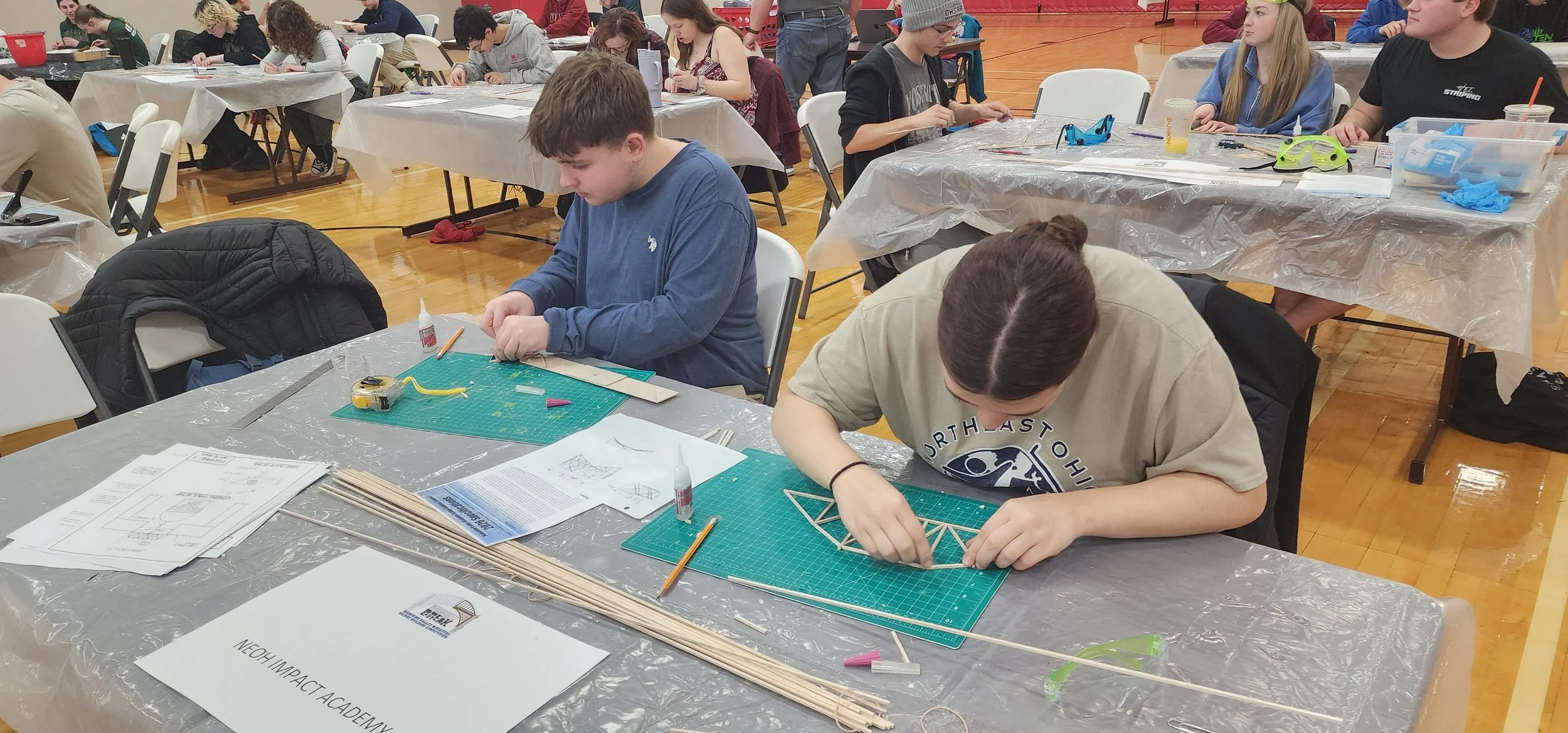 Students working on model construction projects at tables covered with plastic sheets in a gymnasium.