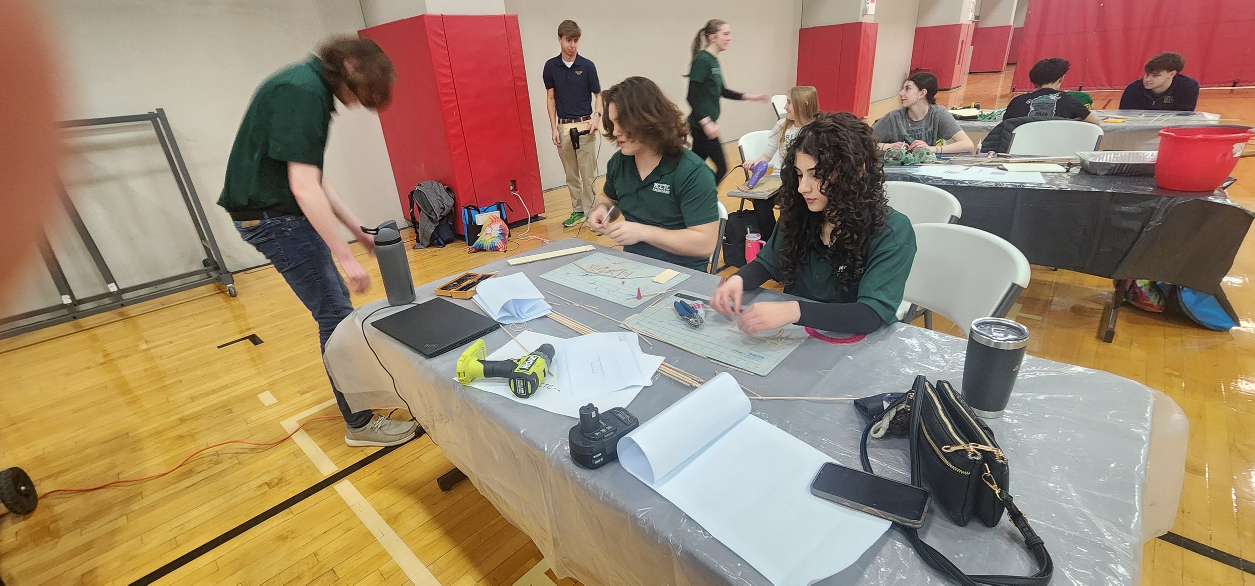 Group of students working on projects at a table in a gymnasium, with tools and materials scattered on the table, and bags and equipment in the background.