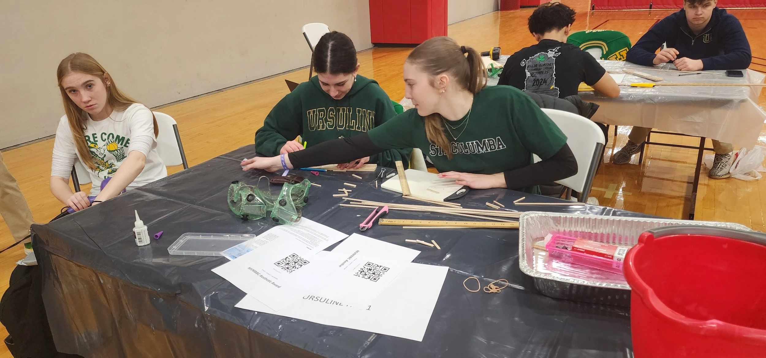Students working on a project at a table covered with crafting supplies, rulers, sticks, and tools in a gymnasium.