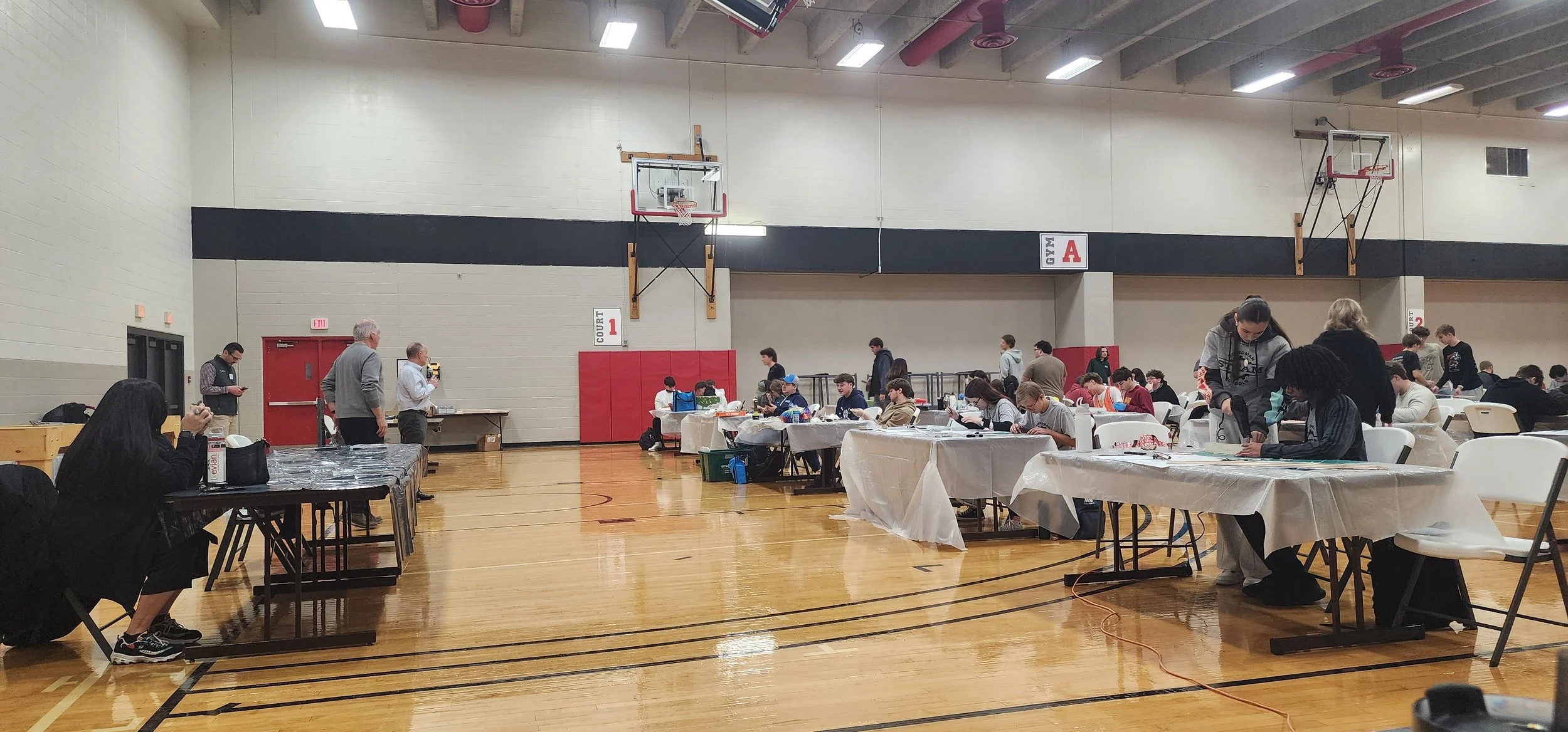 A gymnasium filled with people sitting at tables covered with white and black tablecloths, engaged in various activities. The gym has basketball hoops on the walls, and the floor is made of wood. Several individuals are standing, walking, or working 