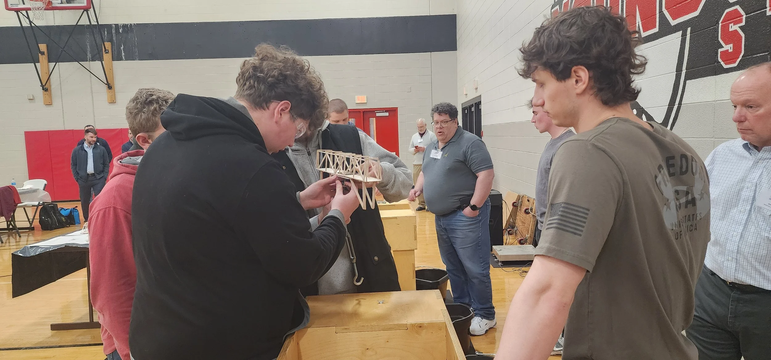 A group of young men and adults gather around a table in a gymnasium, inspecting a small wooden model of a bridge.
