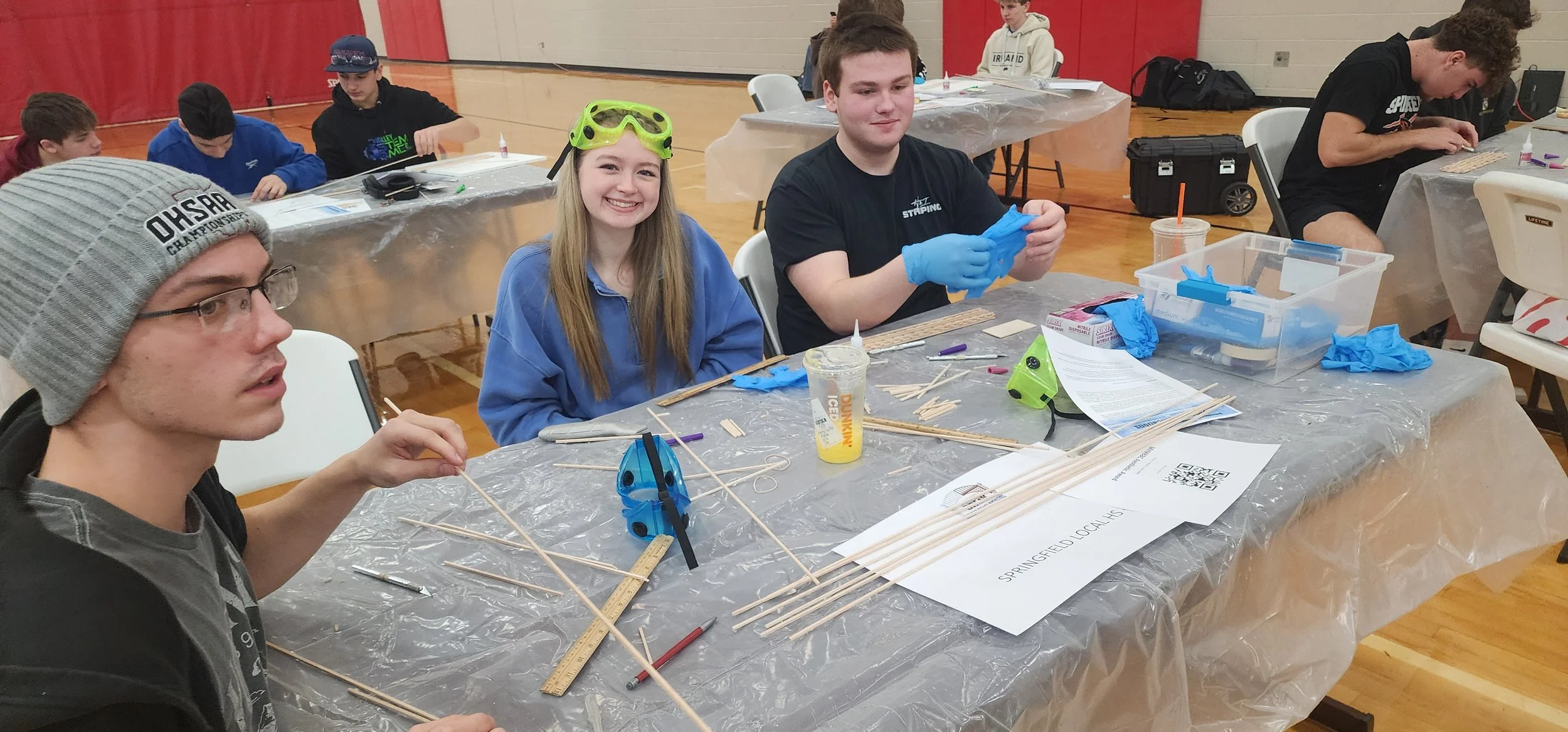 Group of young people sitting at a table working on a craft project with wooden sticks, rulers, glue, and scissors, in a gymnasium.