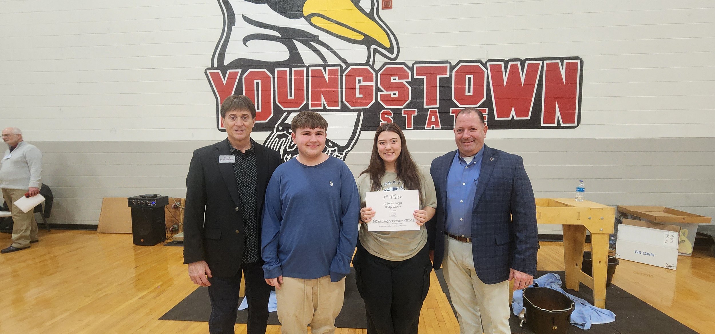 A group of four people standing in front of a large Youngstown State logo painted on a gymnasium wall, with one person holding a first-place certificate at an award event.
