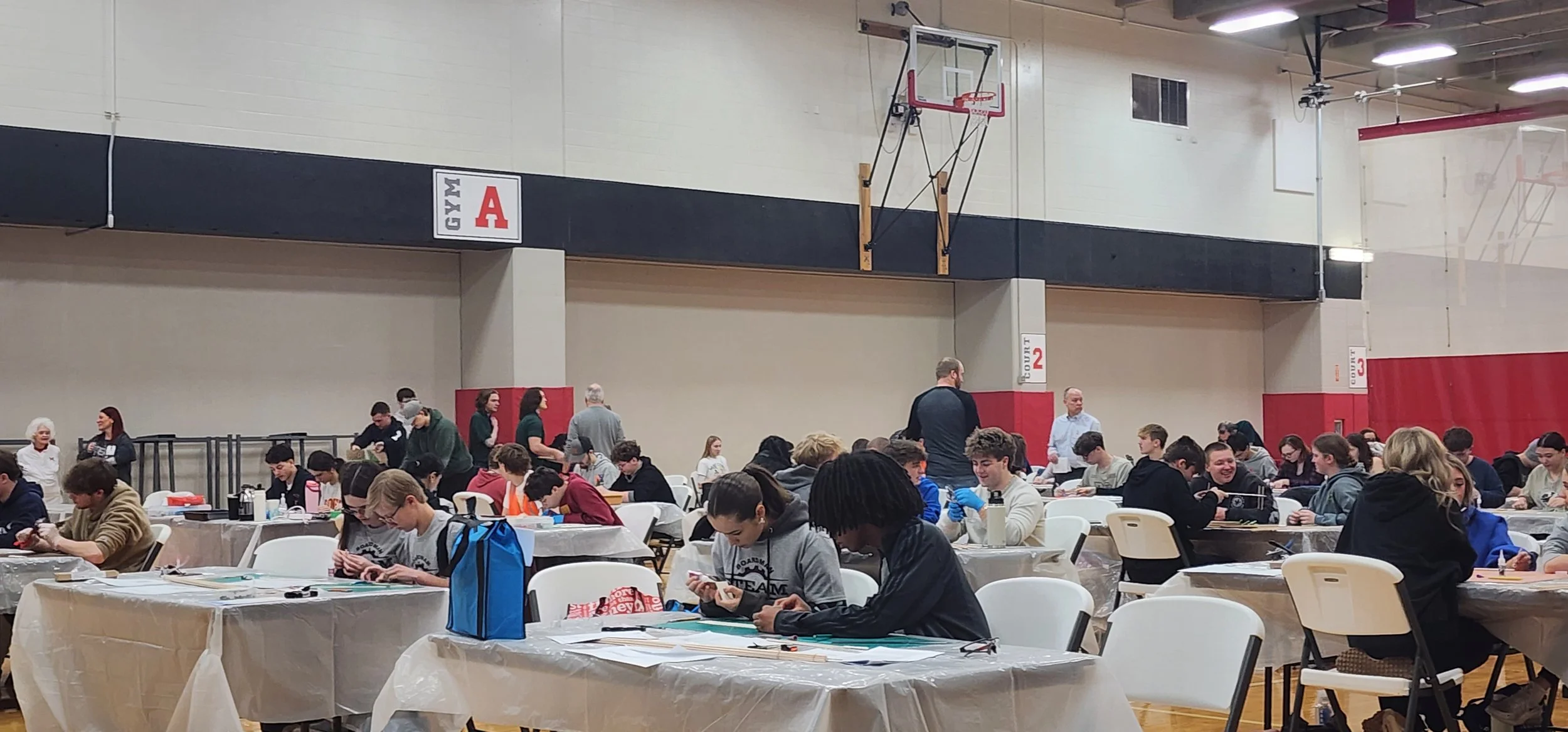 Students participating in a workshop or class inside a gymnasium, working on individual projects at tables covered with plastic, with teachers and monitors assisting.
