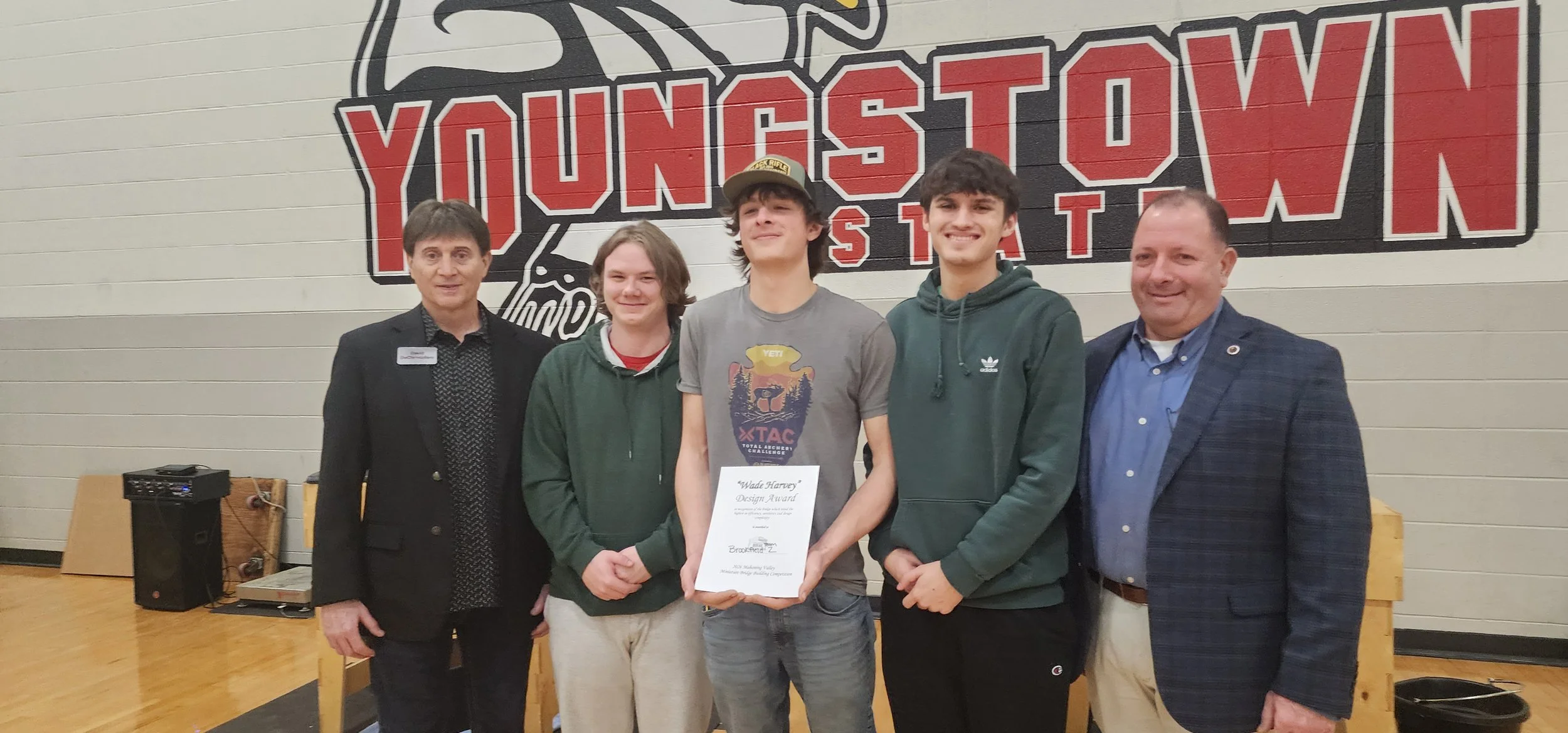 A group of five people standing in front of a wall with a large logo that says 'Youngstown State University' and a mountain lion mascot. The group includes three young men and two older men, one of whom is holding a certificate.