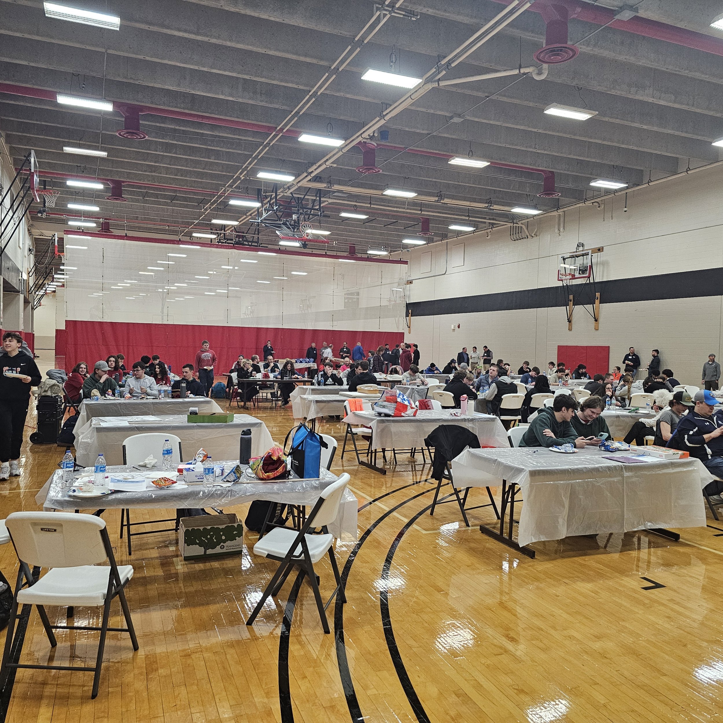 A gymnasium filled with people sitting at tables covered with plastic tablecloths, eating and socializing. The gym has a high ceiling with bright lights, basketball hoops, and red accent colors on the walls and ceiling.