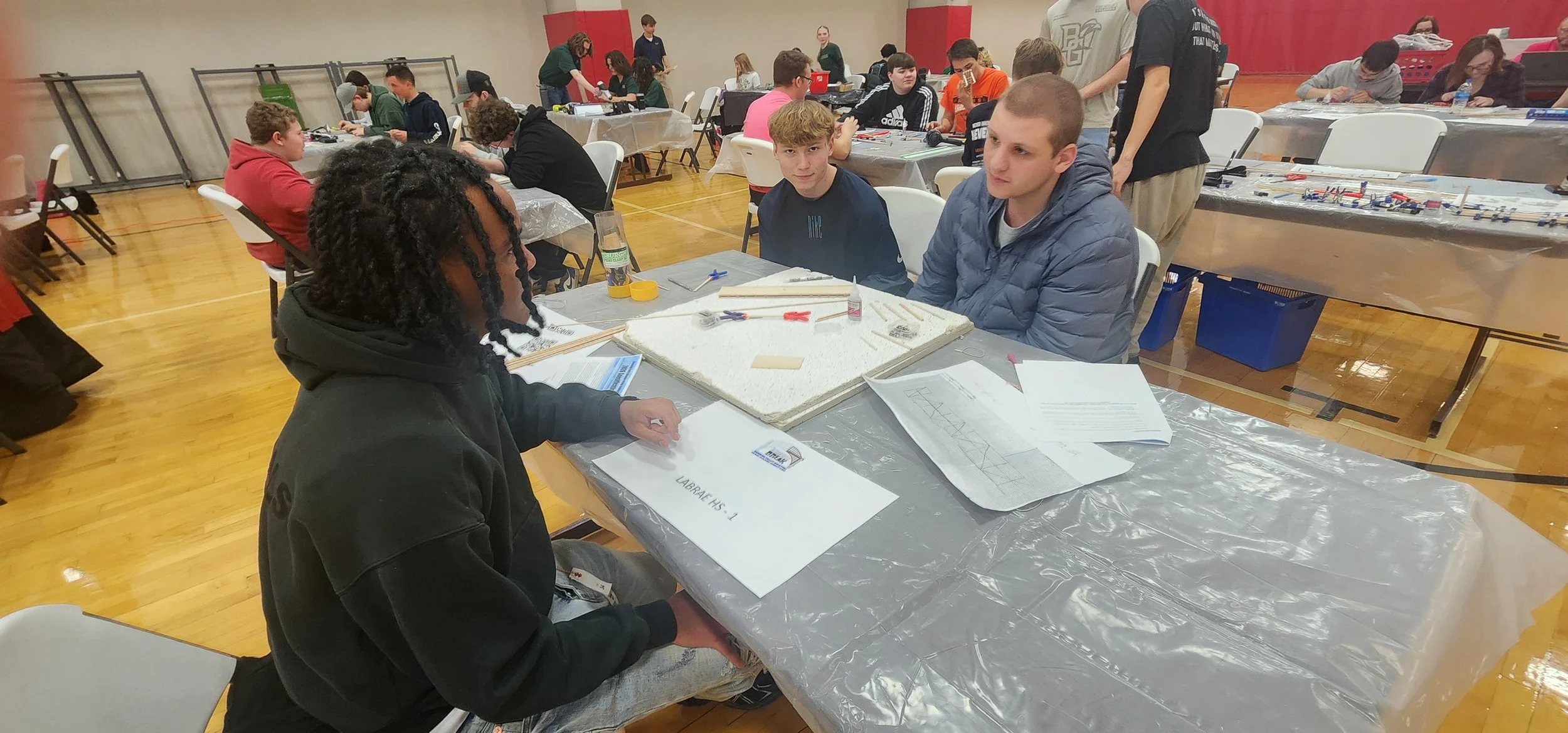 Several students seated around tables working on robotics projects in a gymnasium, with various tools and papers scattered on the tables.