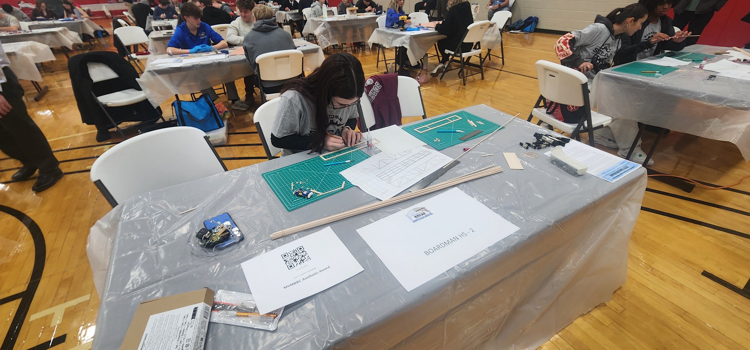 A girl working on a model at a table covered with a plastic tablecloth in a gymnasium filled with other students working on projects.