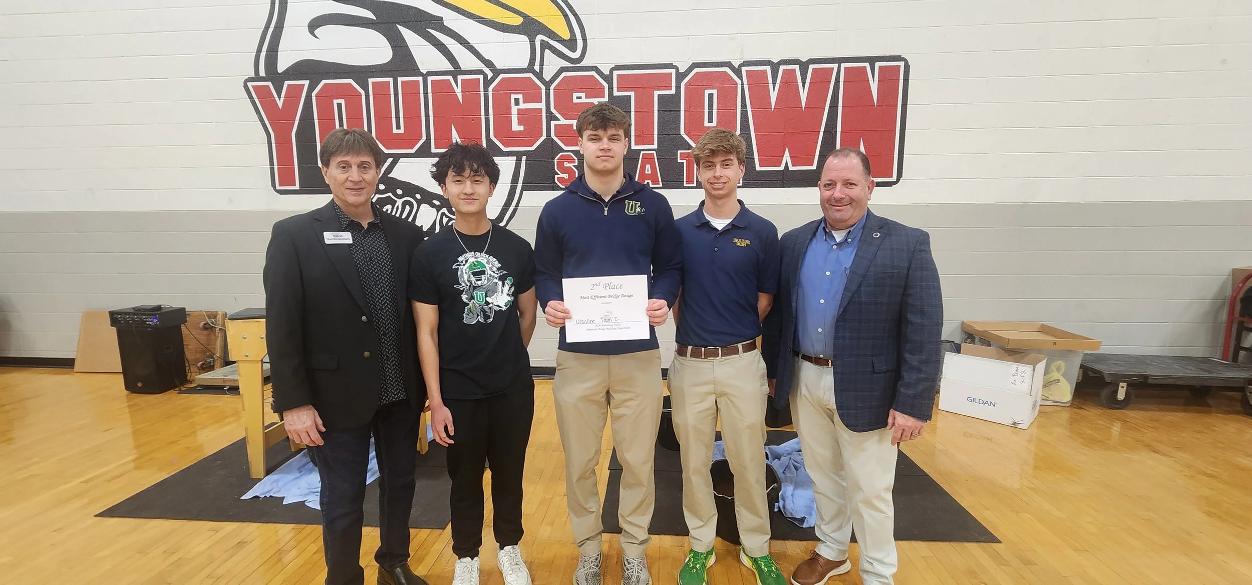 Group of five people standing in front of a large 'Youngstown State' sign, three young men holding a second-place award for 'Most Efficient Bridge Design,' flanked by two adult men.