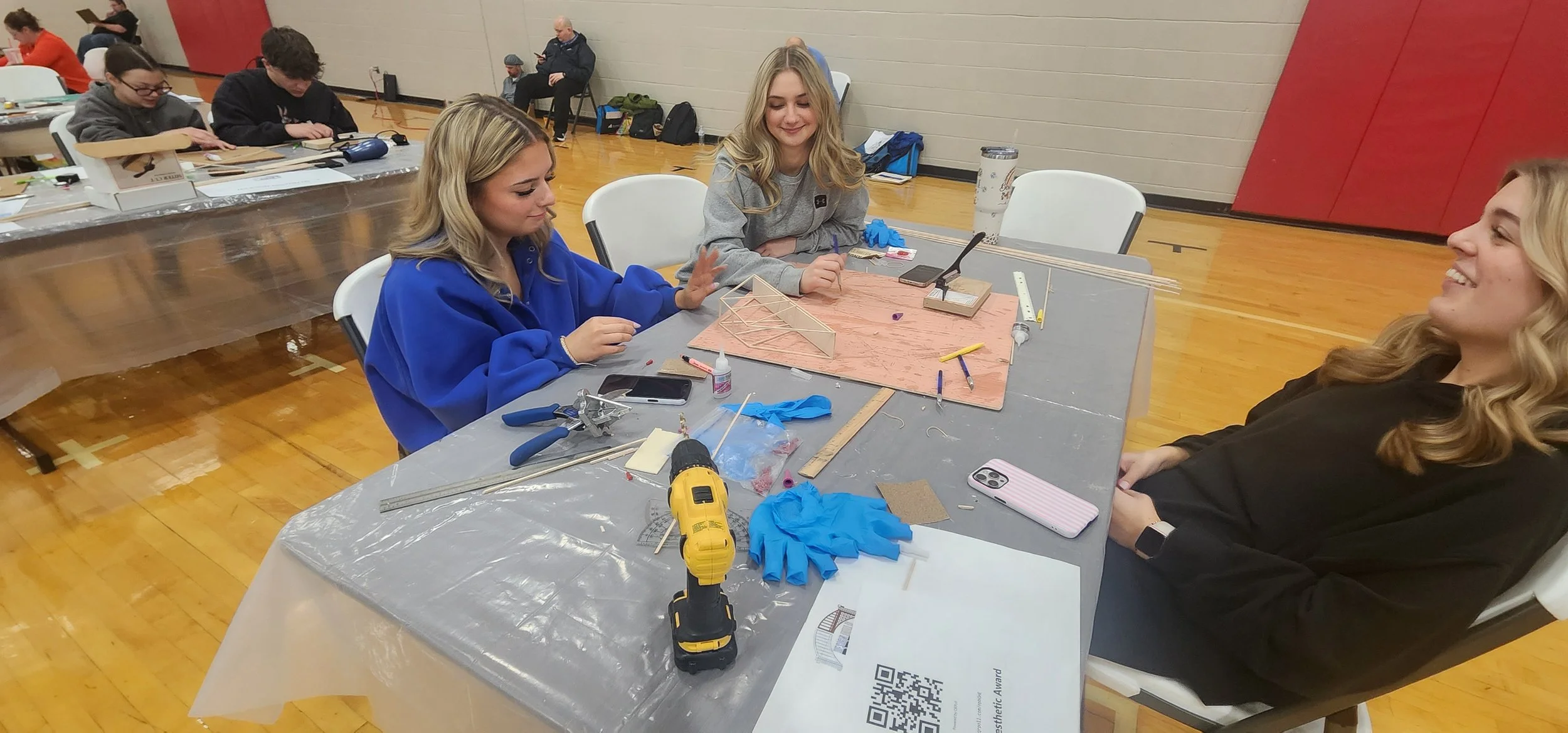 Three young women sitting at a table working on a project, with tools and materials scattered on the table; two are smiling and engaging with each other, and one is gesturing with her hand.