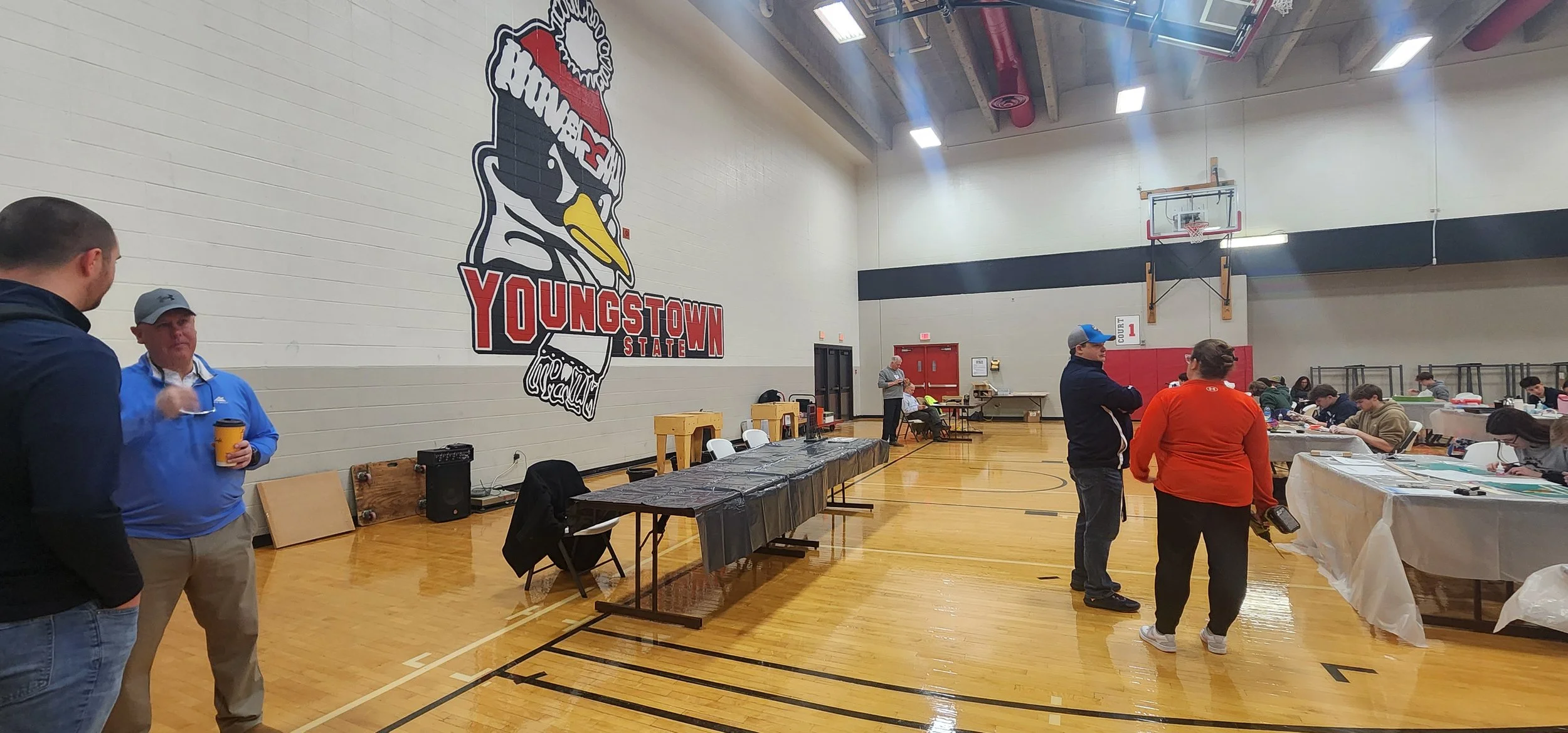 Inside a gymnasium at Youngstown State, people are gathered around tables, some engaged in conversations, with a large Youngstown State logo on the wall and a basketball hoop in the background.
