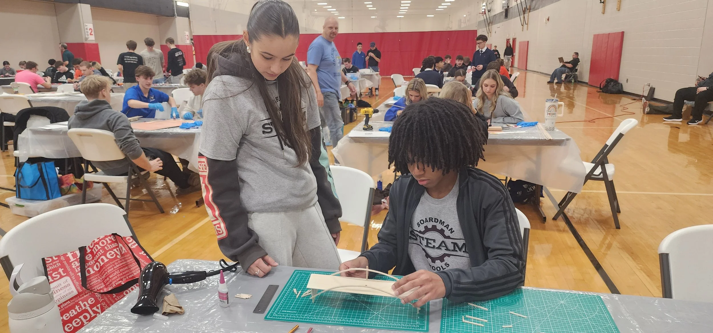 At a robotics workshop, a young woman with long brown hair looks down at a seated young man with dreadlocks working on a project. The room is filled with other young participants and some adults, all engaged in various activities at tables in the bac