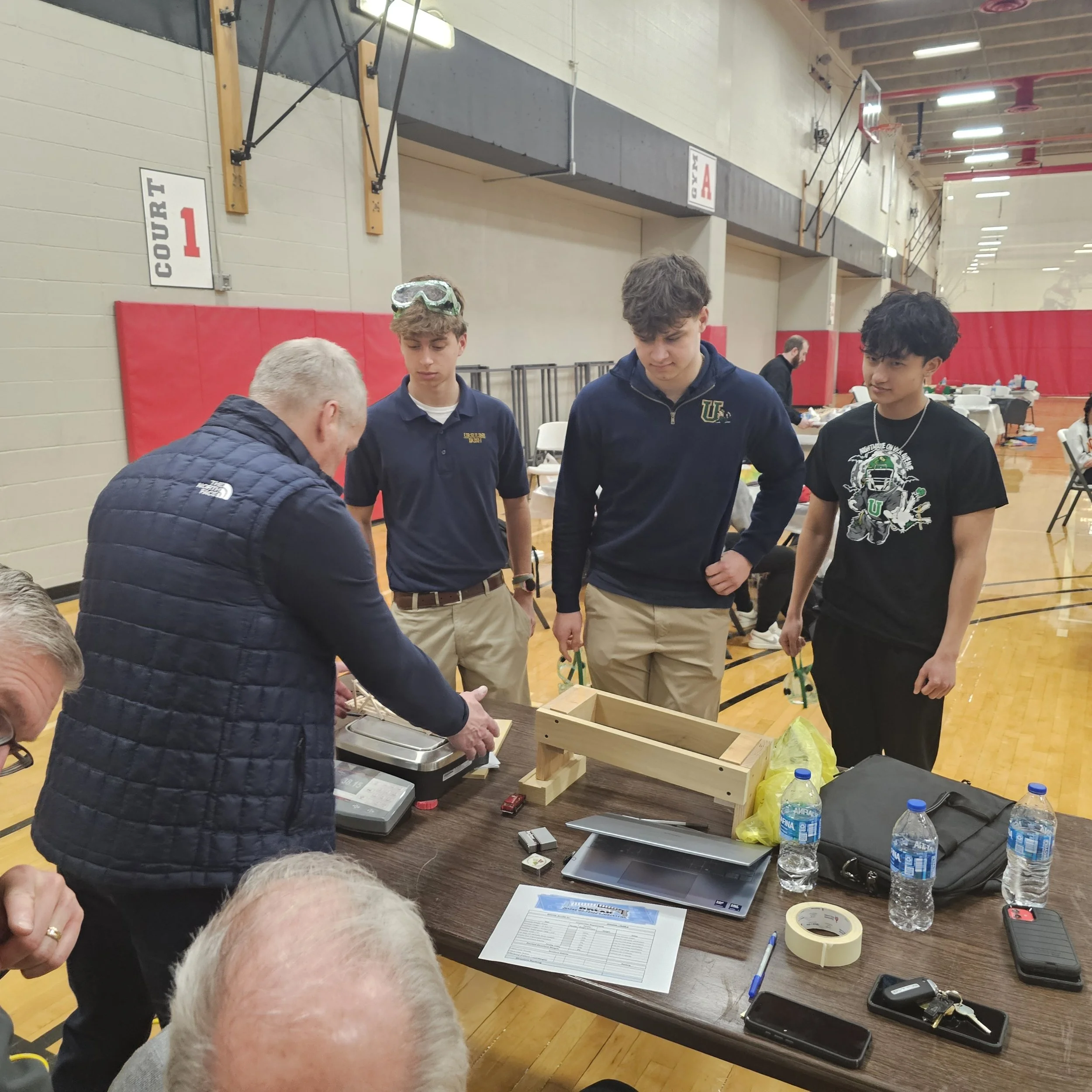 Four young men stand around a table with water bottles, paper, and various objects, possibly working on a project or experiment inside a gymnasium. Two are wearing dark hoodies, one with goggles on his forehead, and one has a graphic t-shirt. A man w