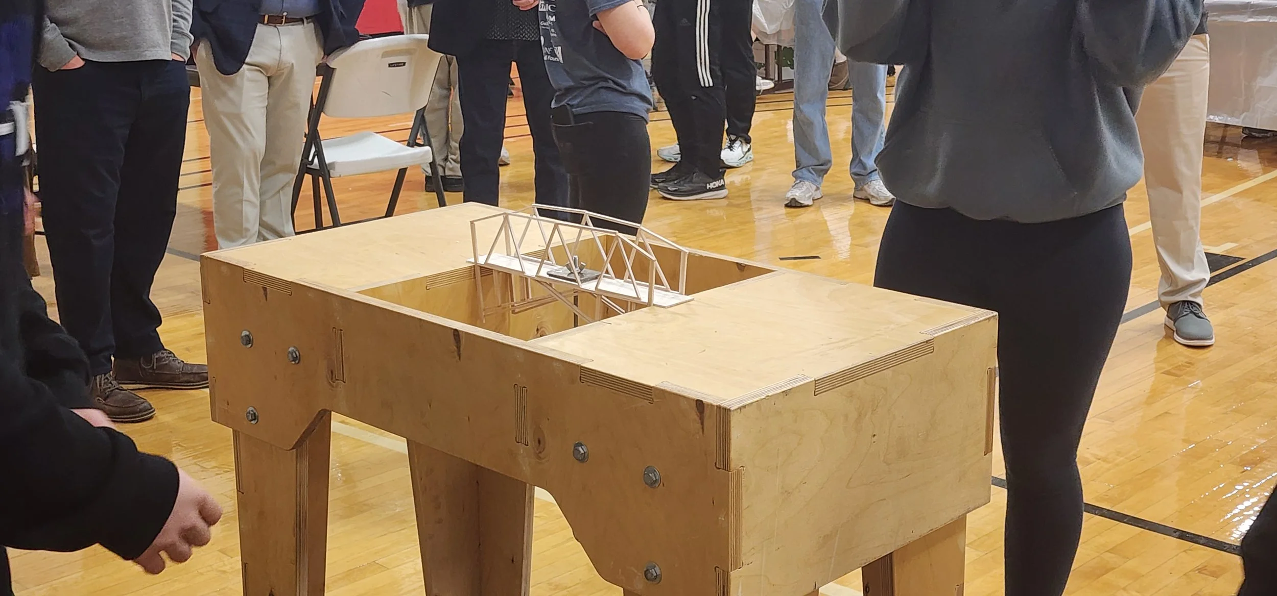Group of people gathered around a wooden table with a model of a bridge on top in a gymnasium.