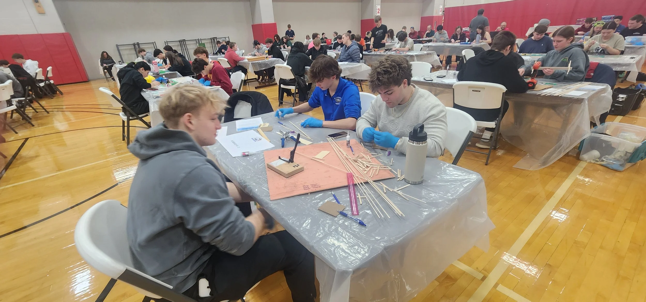 Students working on a woodworking project at long tables covered with plastic, with tools, wood pieces, and measurement equipment. A gymnasium with a wooden floor and red padded walls.