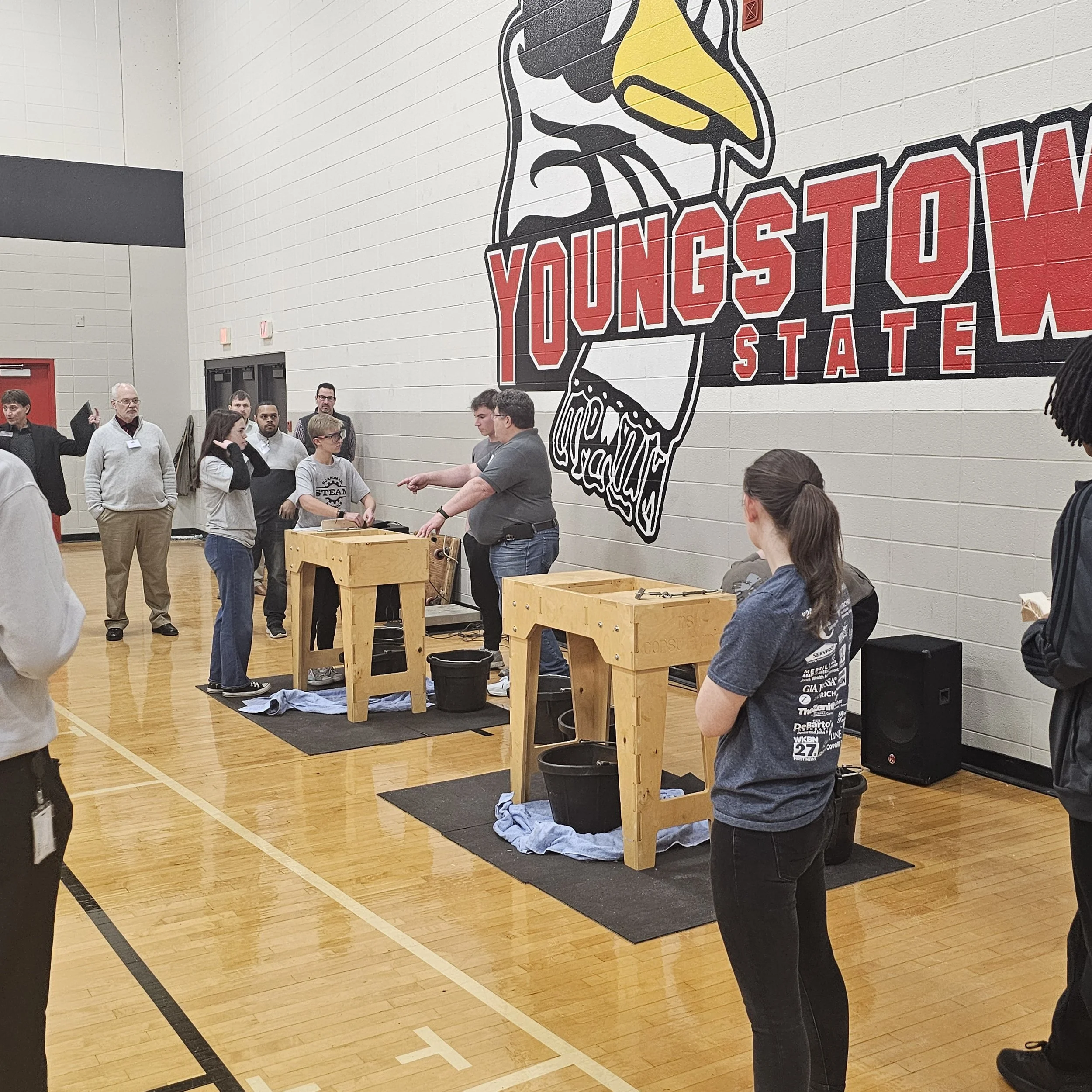 People gathered in a gymnasium with a large artwork of the state of Ohio and the words "Youngstown State" displayed on the wall.