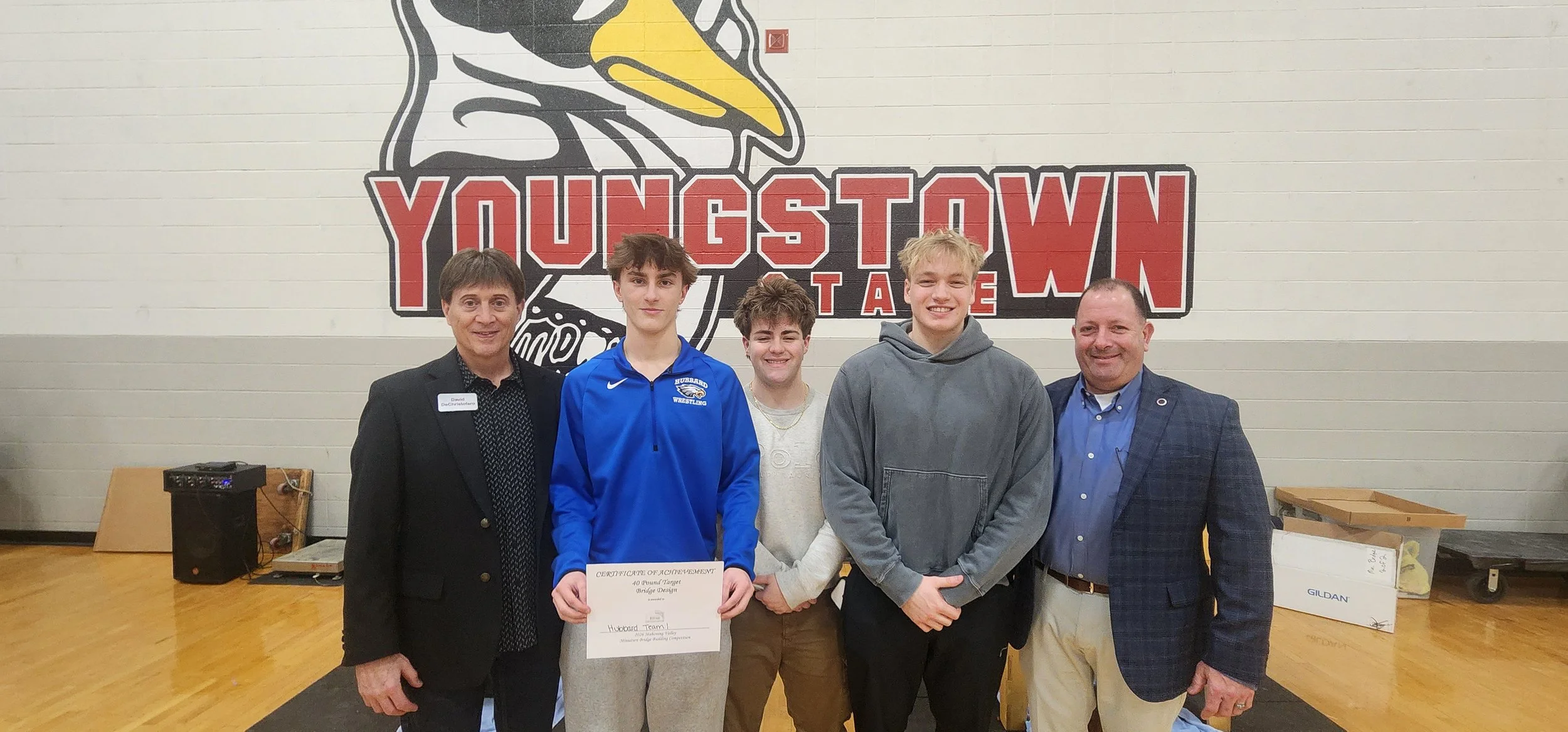 Group of five people standing in front of a large logo that reads 'Youngstown' with a stork head illustration. One person in the center holds a certificate. The setting appears to be an indoor event or awards ceremony.