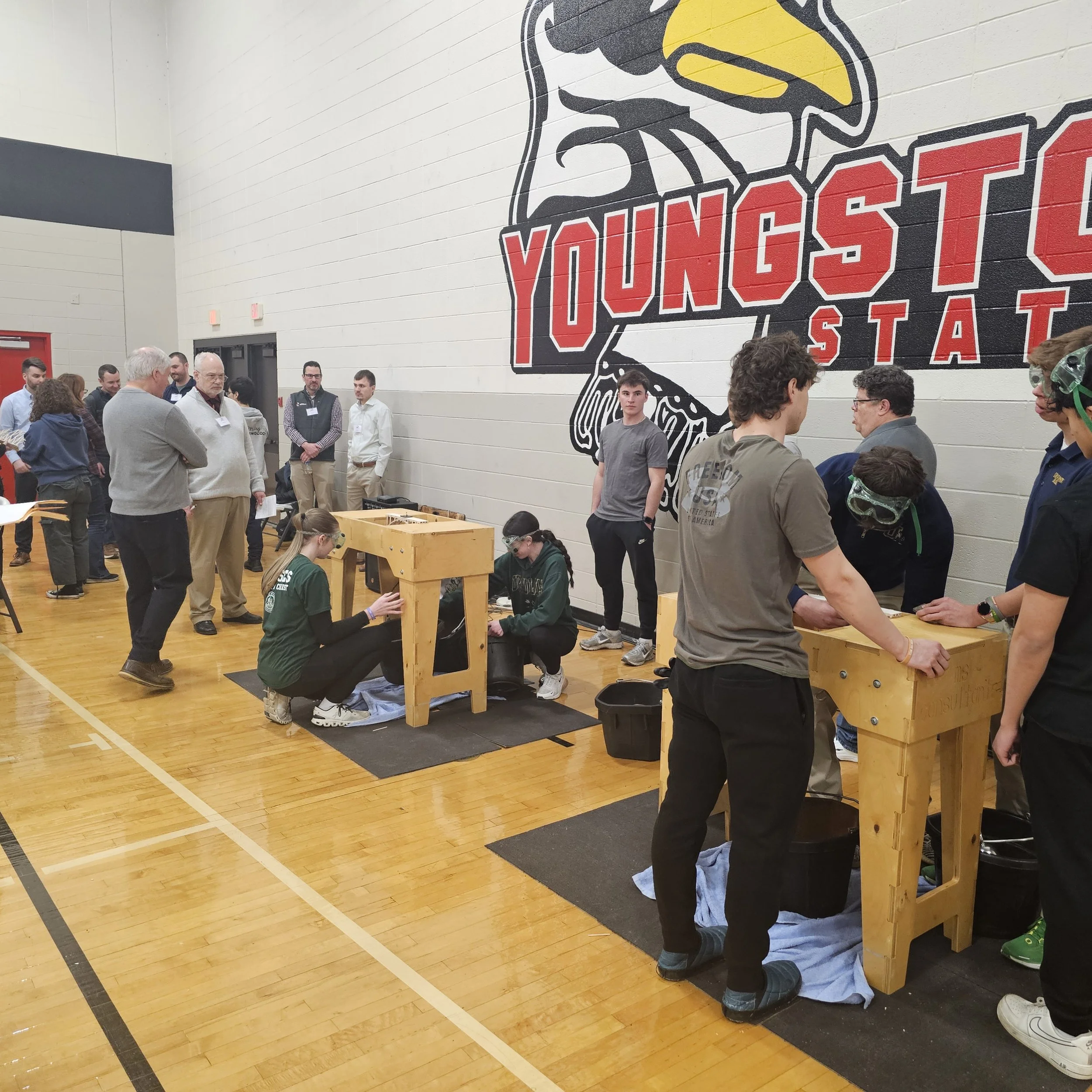 Group of people gathered indoors, some standing and others kneeling, around wooden workbenches. Large sports logo or mascot of a bird with a yellow beak painted on the wall.