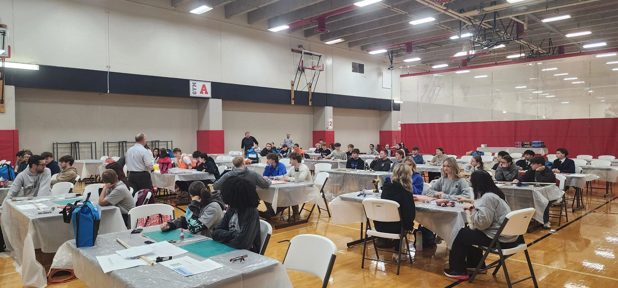 Students seated at tables in a gymnasium, engaged in a hands-on activity, with some adults overseeing. The gym has basketball hoops and a sign that says 'GYM A'.