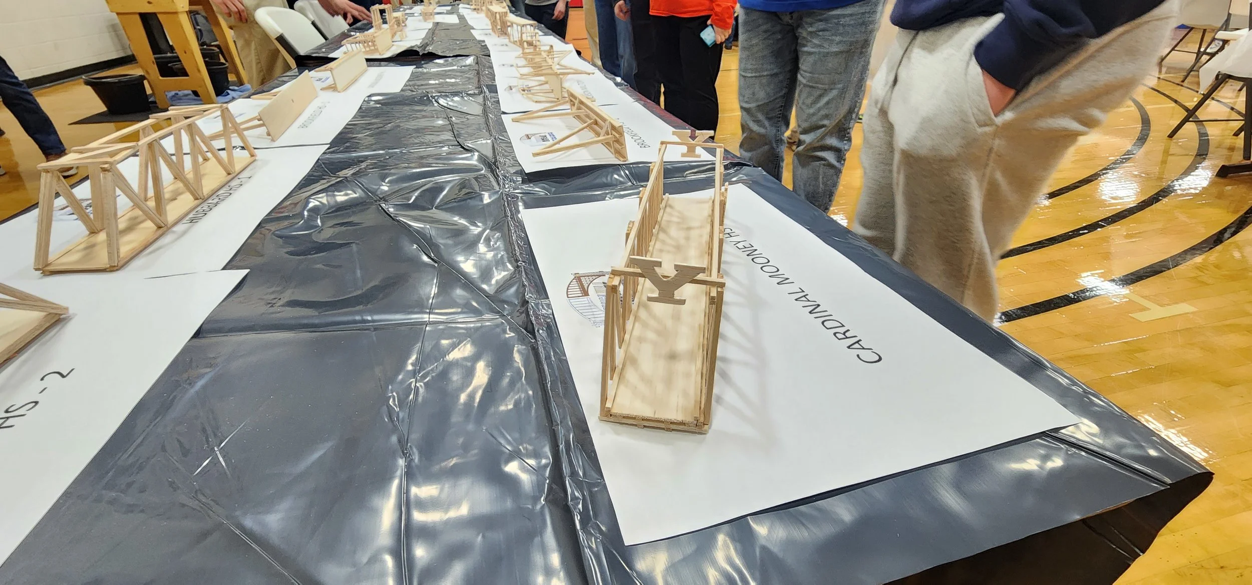 Miniature wooden bridges and structures displayed on a table at an indoor event, with people observing.