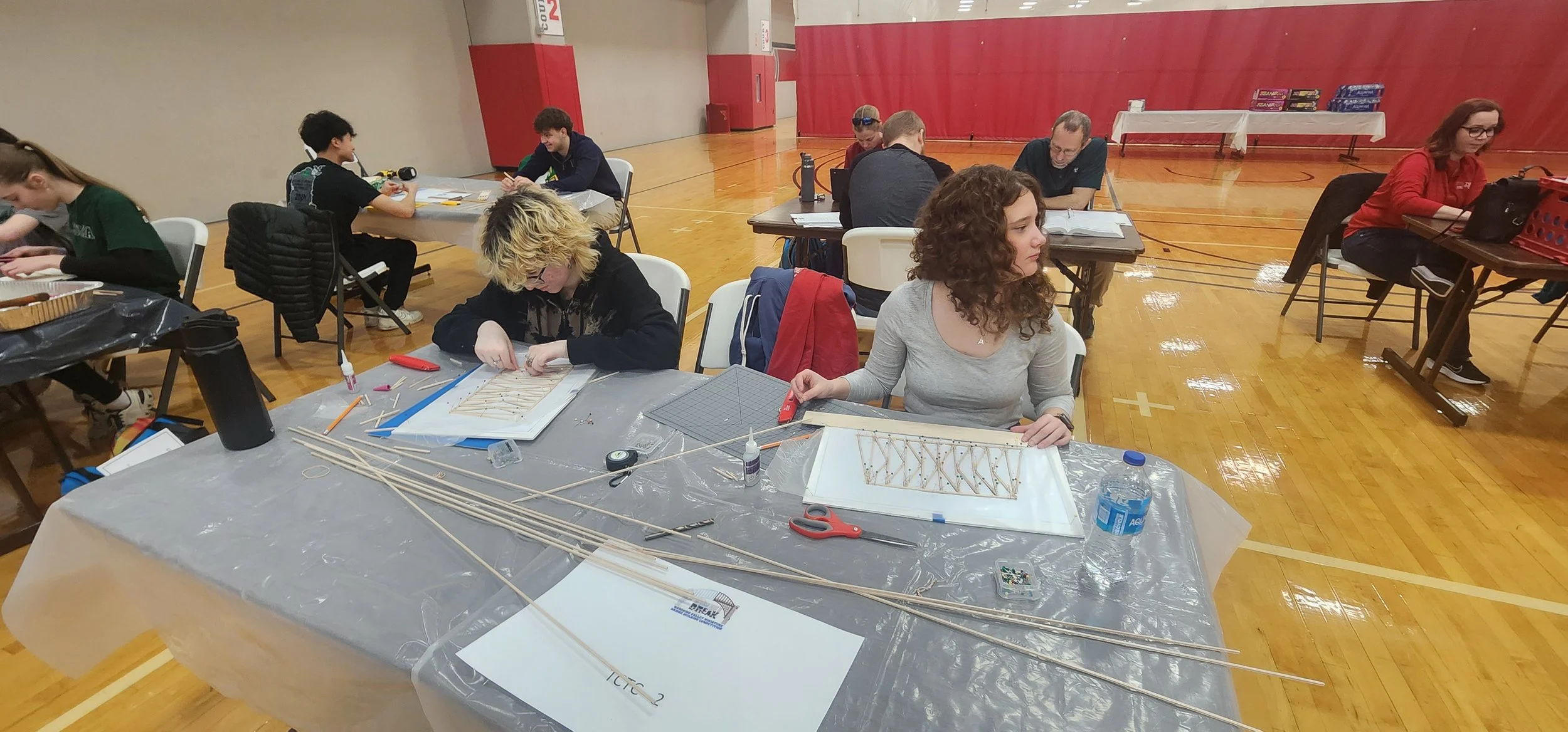 People working on model bridges at tables in a gymnasium with wooden floors and red walls.