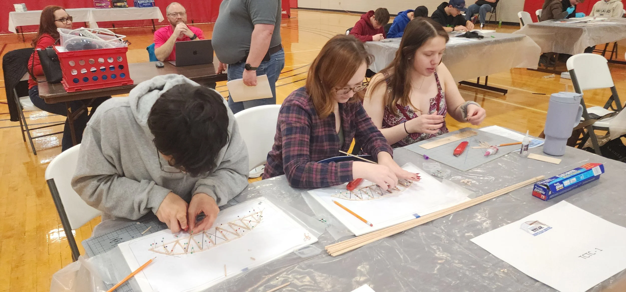 People working on arts and crafts projects at a table set up in a gymnasium, with various craft supplies including sticks, glue, and string.