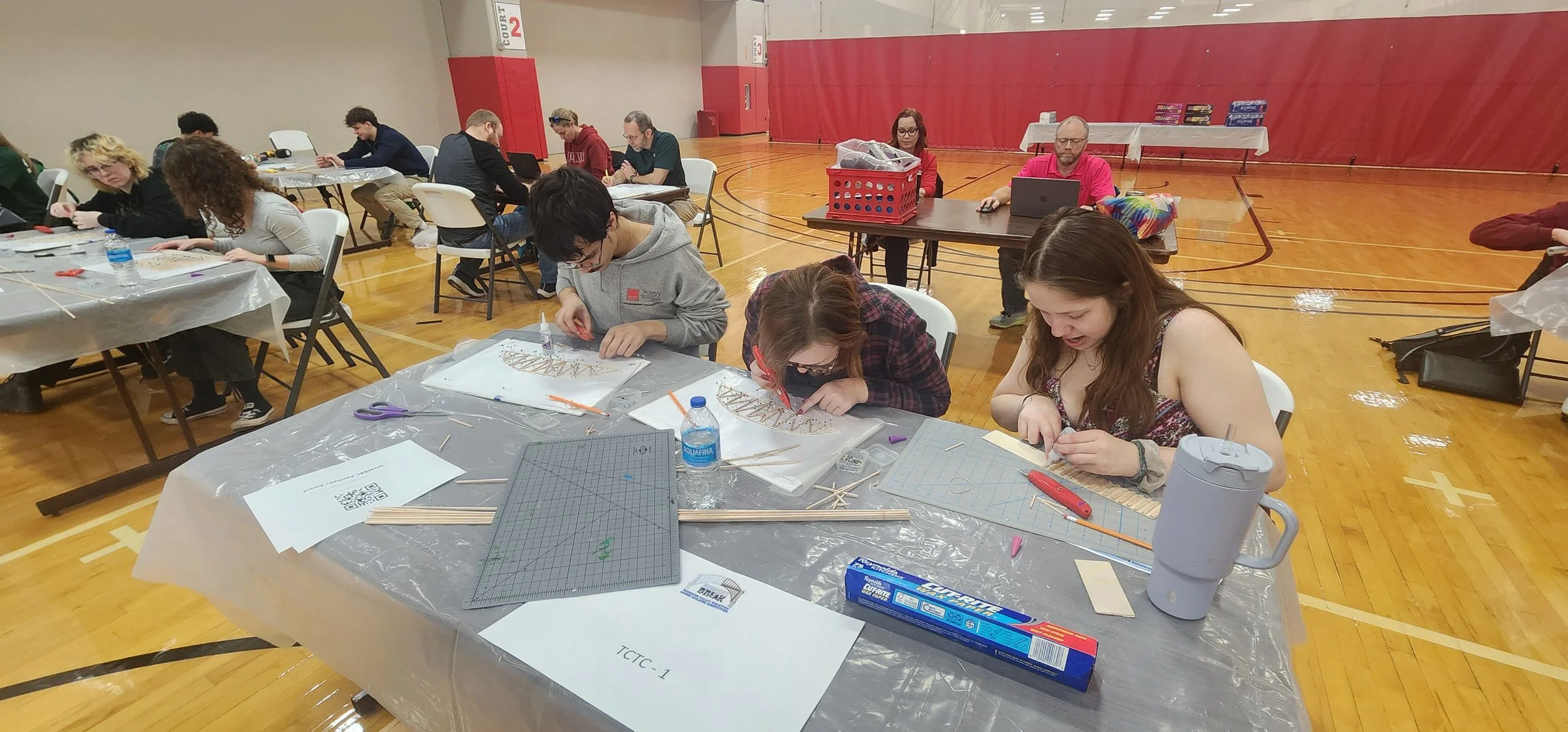 People sitting at tables in a gymnasium engaged in crafting or model building activities, with materials like wood sticks, glue, scissors, and cutting mats on the tables.