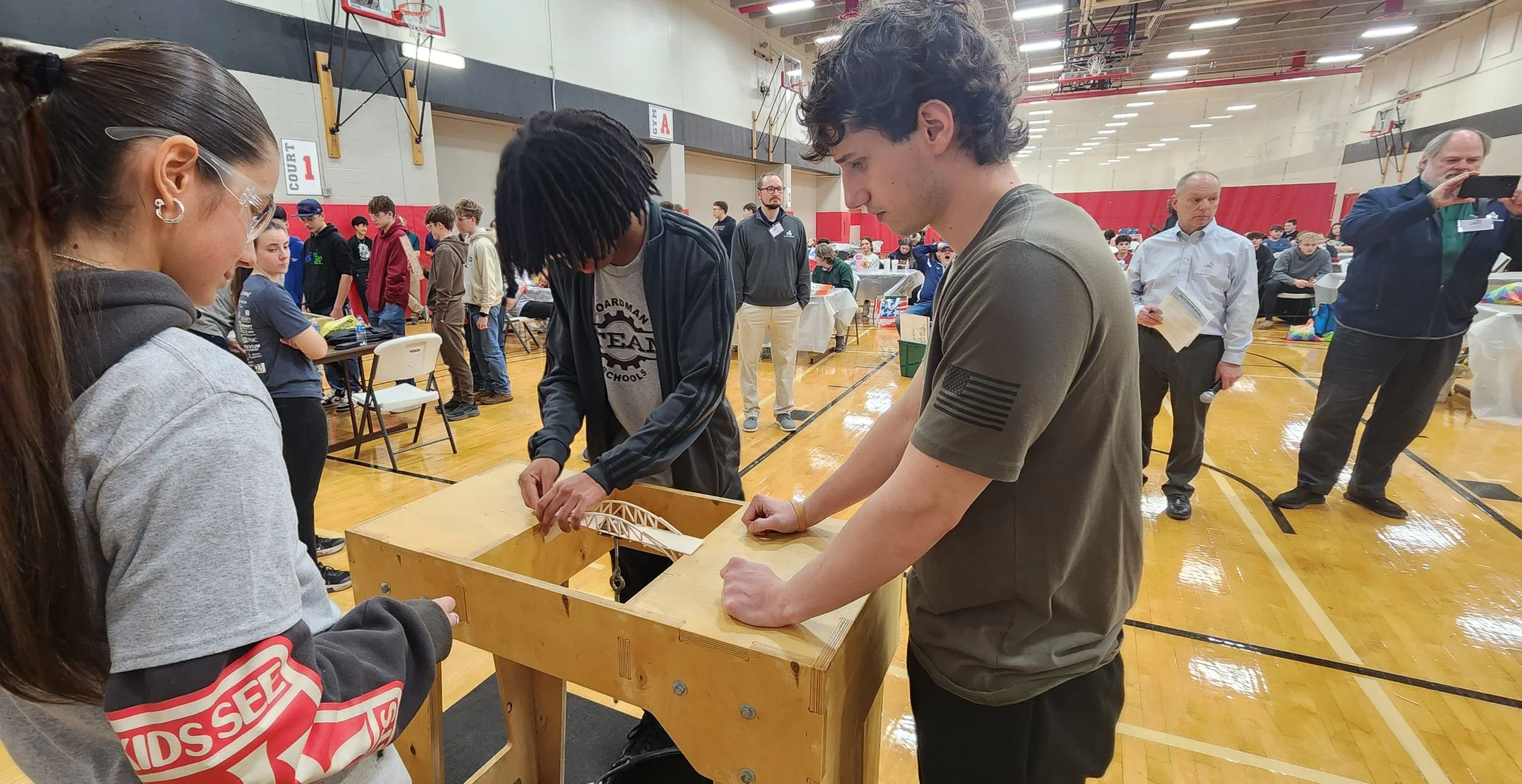 People gathered in a gymnasium around a wooden table, with two young men working on a small bridge model, while others observe and take photos of the activity.