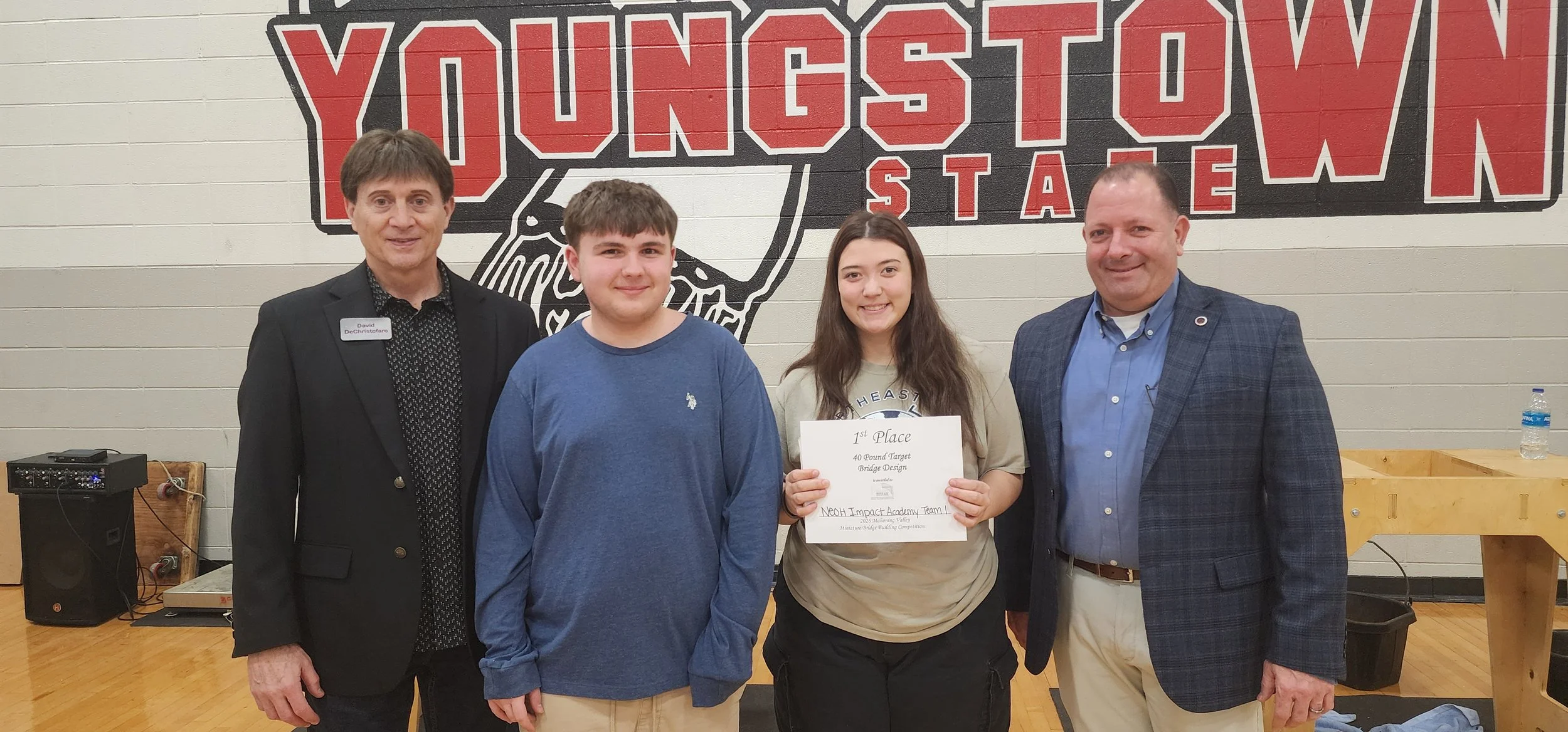Two young students, a boy and a girl, standing between two men in front of a large sign that reads 'YOUNGSTOWN STATE'. The girl is holding a certificate for 1st place in a bridge design competition, and both students are smiling. The setting appears 