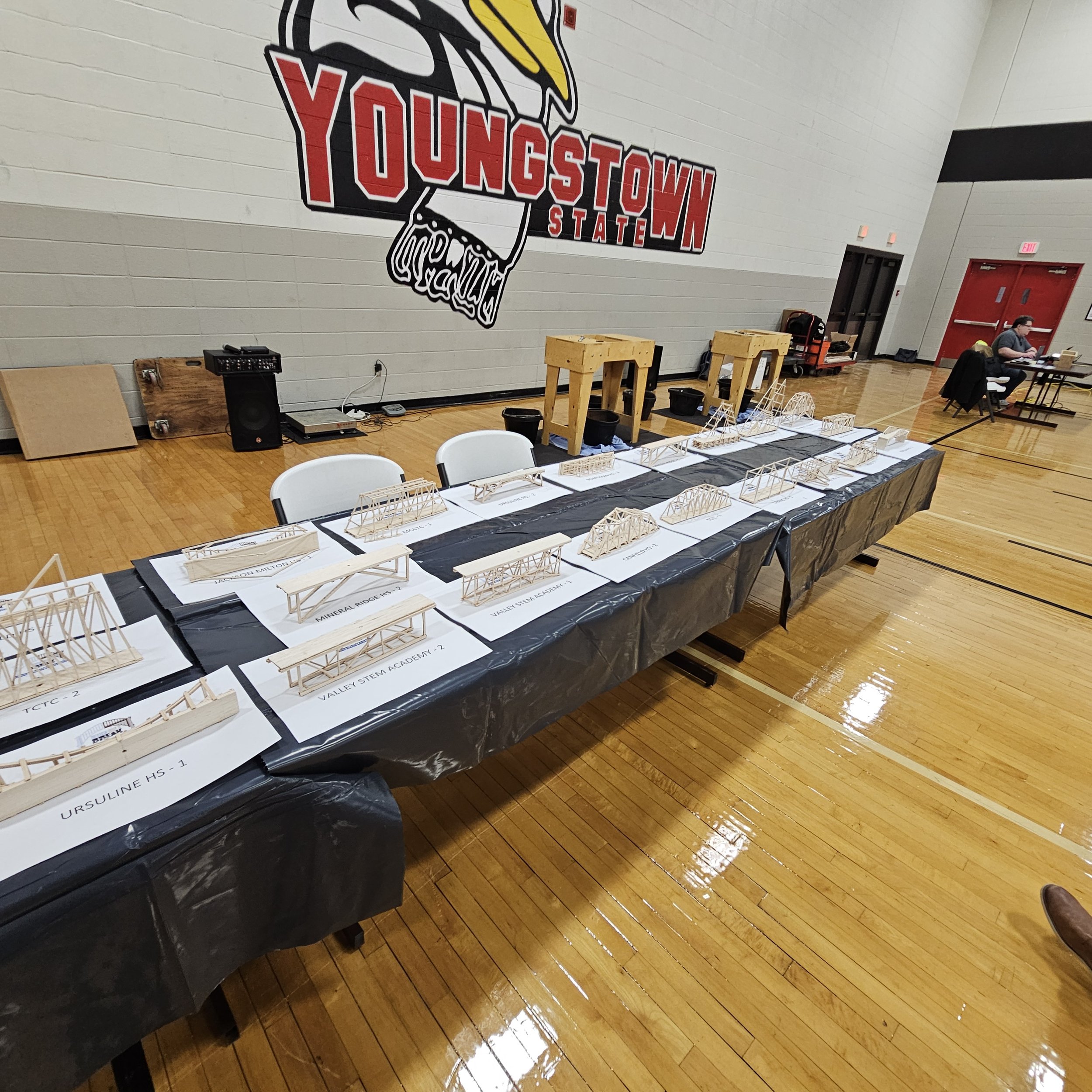 A gymnasium with a large logo on the wall that says 'YOUNGSTOWN STATE' with a bird graphic. There is a table displaying architectural models or miniature structures with labels, and a few people seated at the back. The floor is wooden and there are c