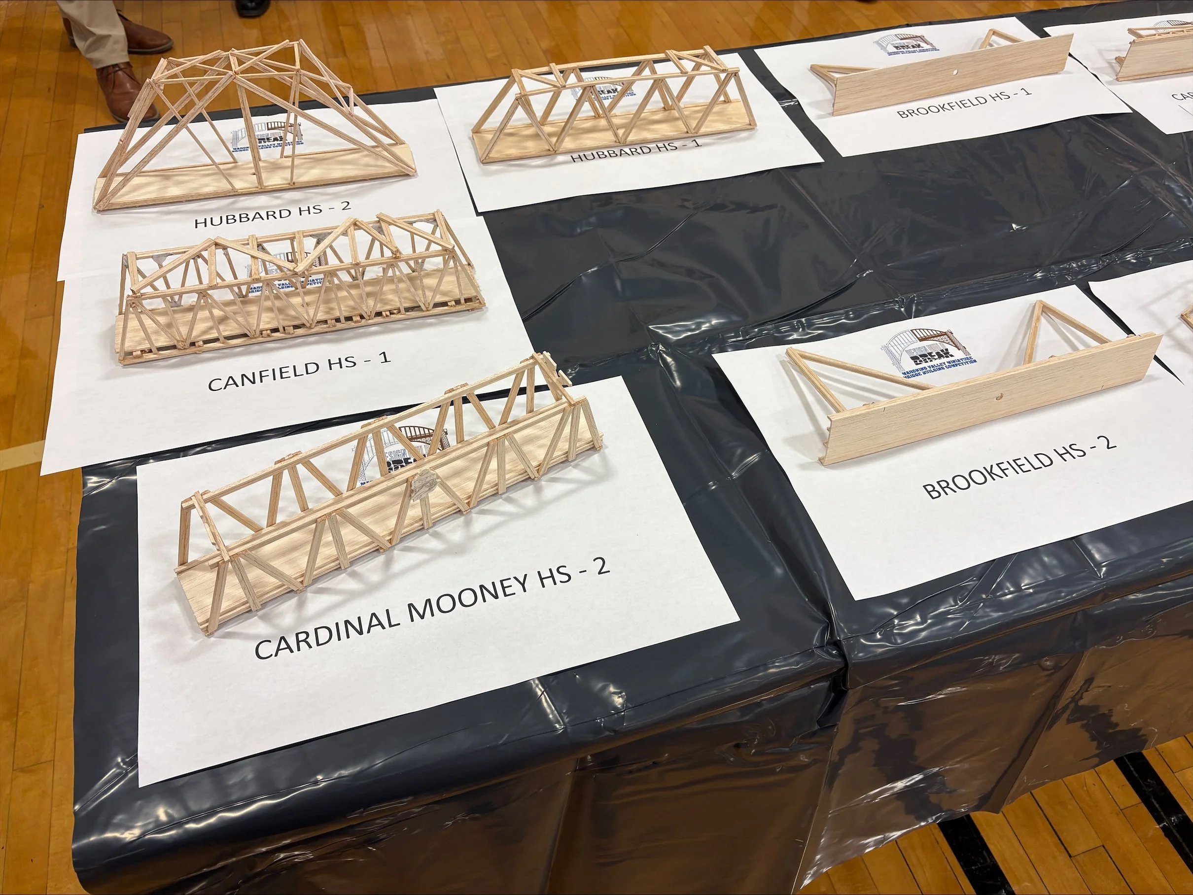 Several wooden model bridges displayed on a table with labels for different high schools. The bridges are constructed from wooden sticks and resemble truss designs.