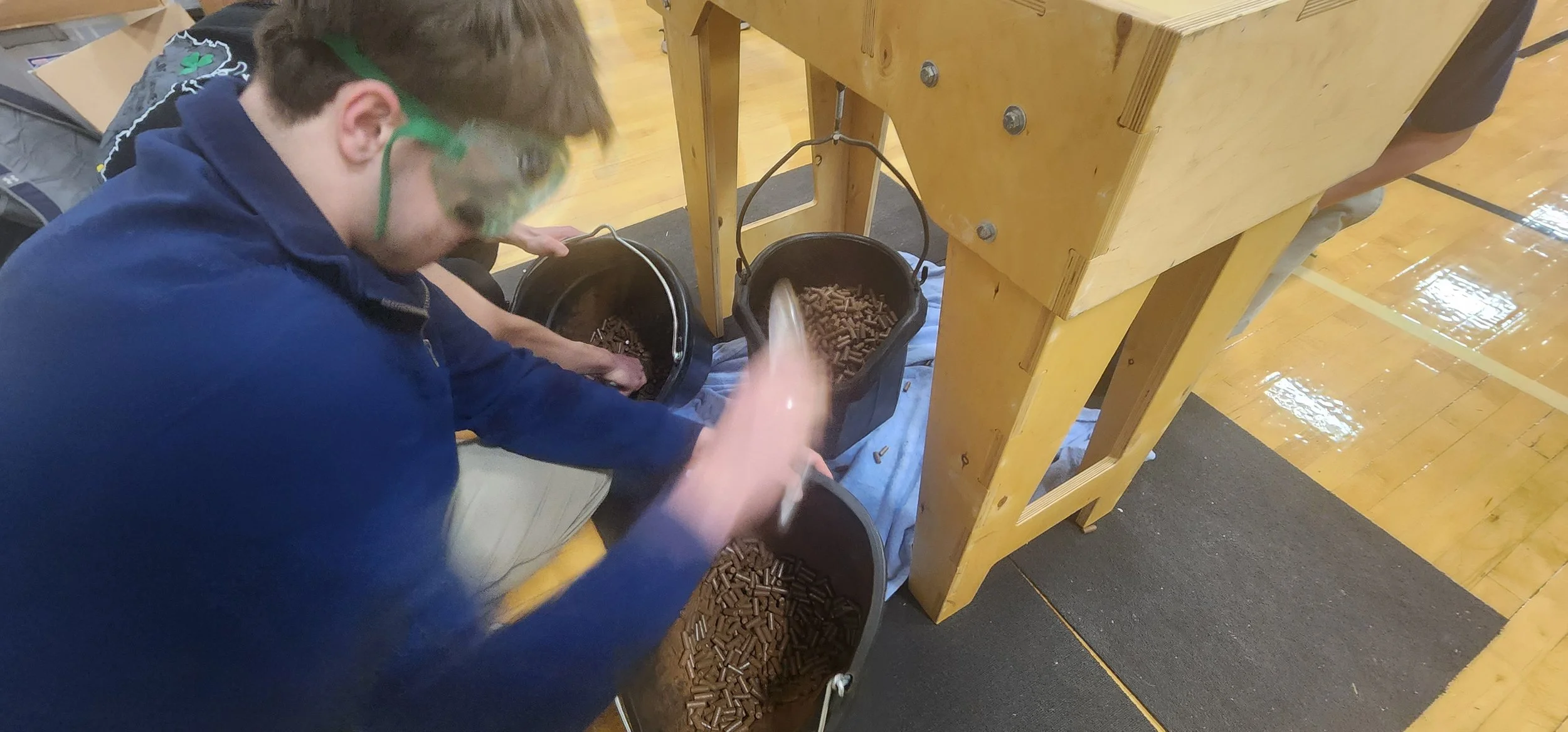 A young person wearing safety goggles and a blue jacket is scooping brown pellets from a black bucket into a smaller container, positioned near a wooden table with other buckets underneath. The setting appears to be an indoor gymnasium with a wooden 