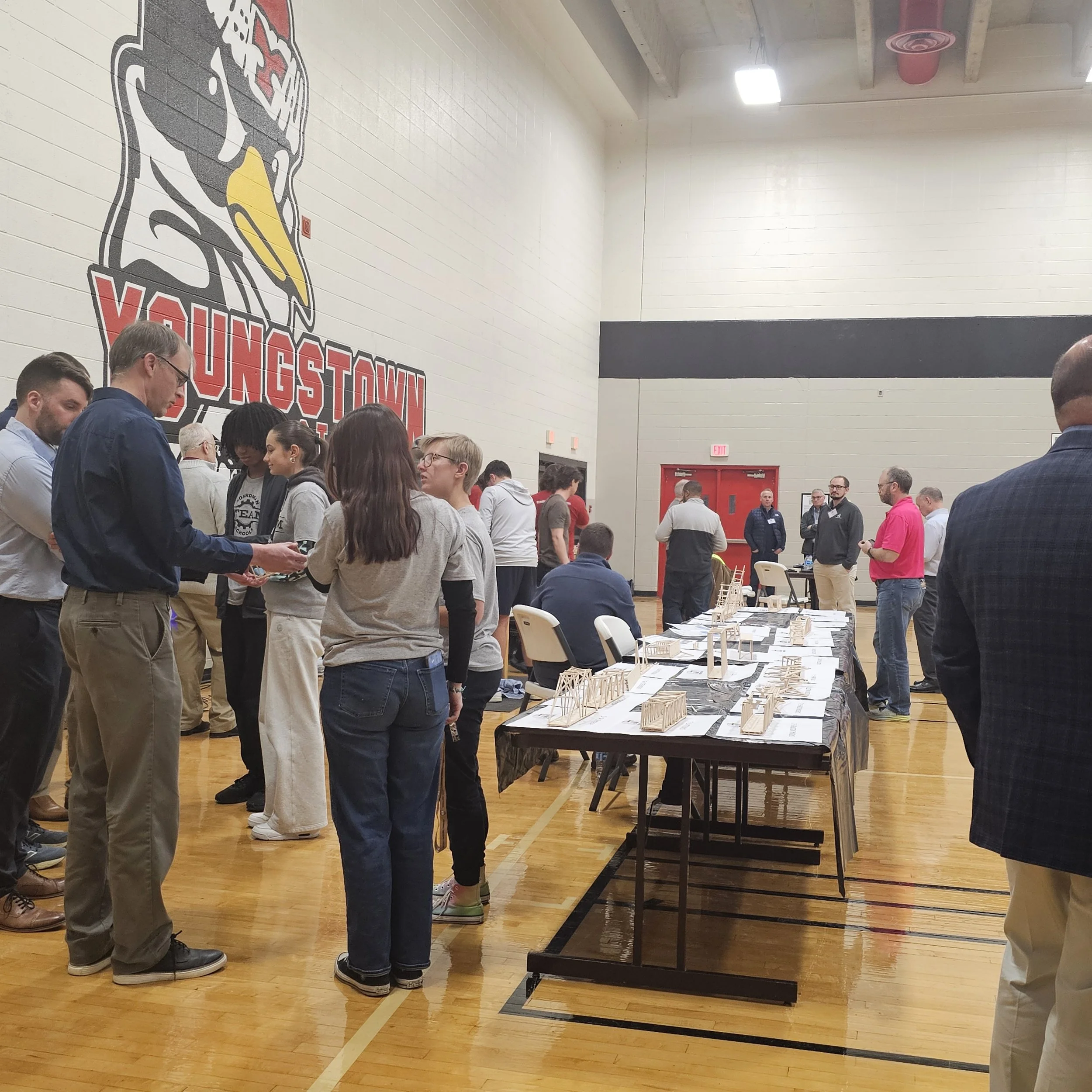 People gather around tables displaying architectural models in a gymnasium, with a large Valkyrie hockey logo on the wall.