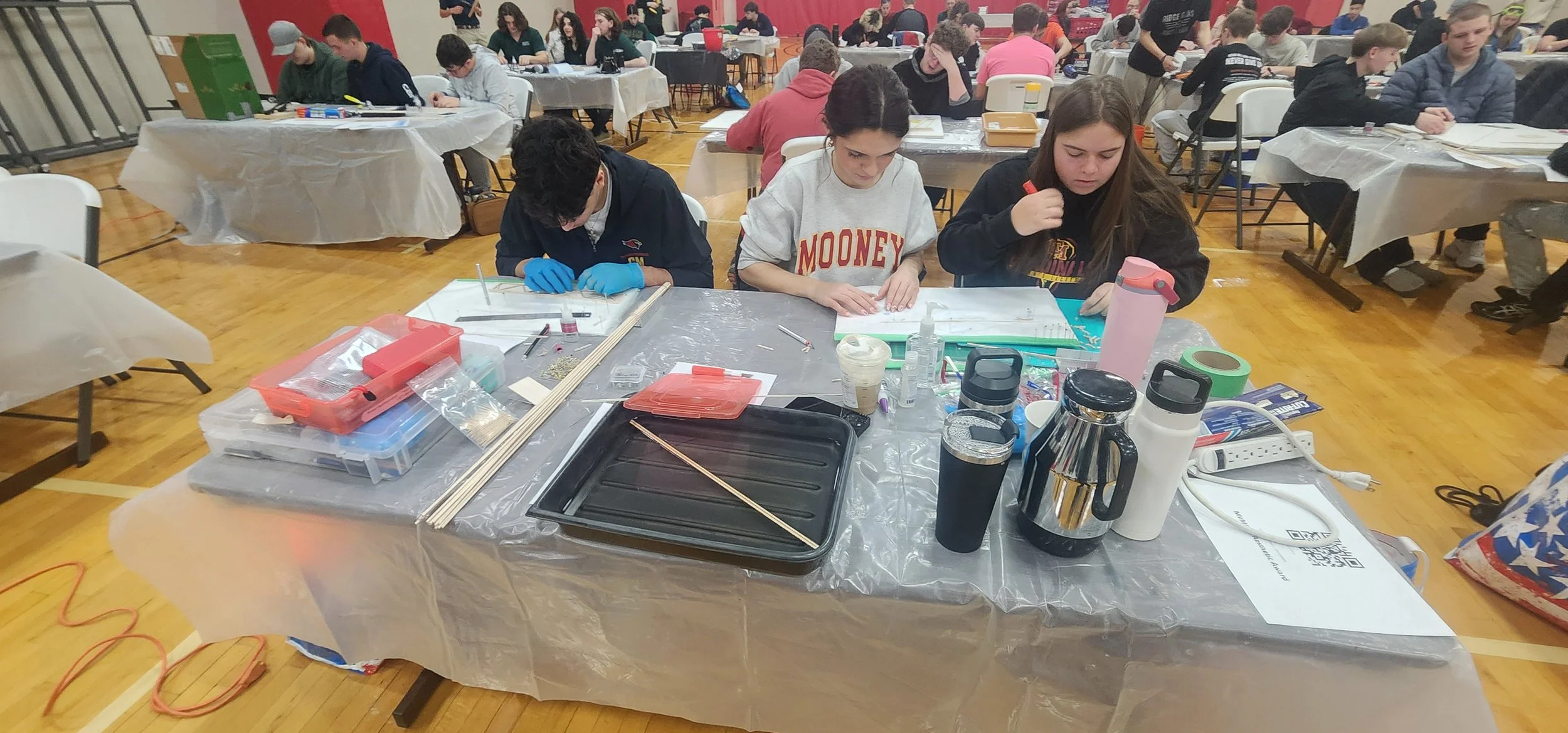 A group of students working on a science or engineering project at long tables covered with plastic tablecloths in a gymnasium. The table has various supplies, tools, and containers. In the background, other students are seated and working at differe