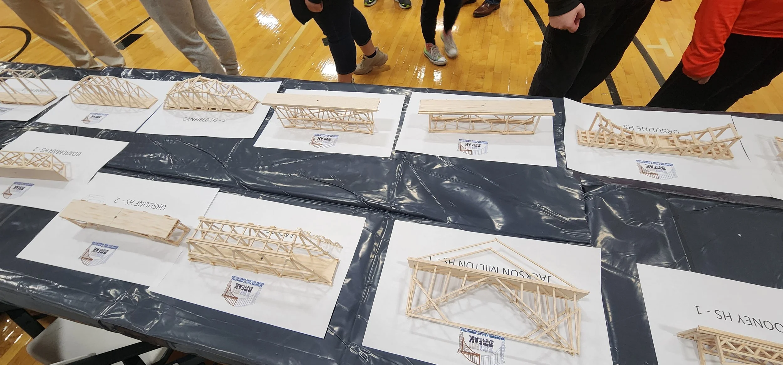 Model bridges made of wooden sticks displayed on white paper sheets on a school gym table, with people observing in the background.