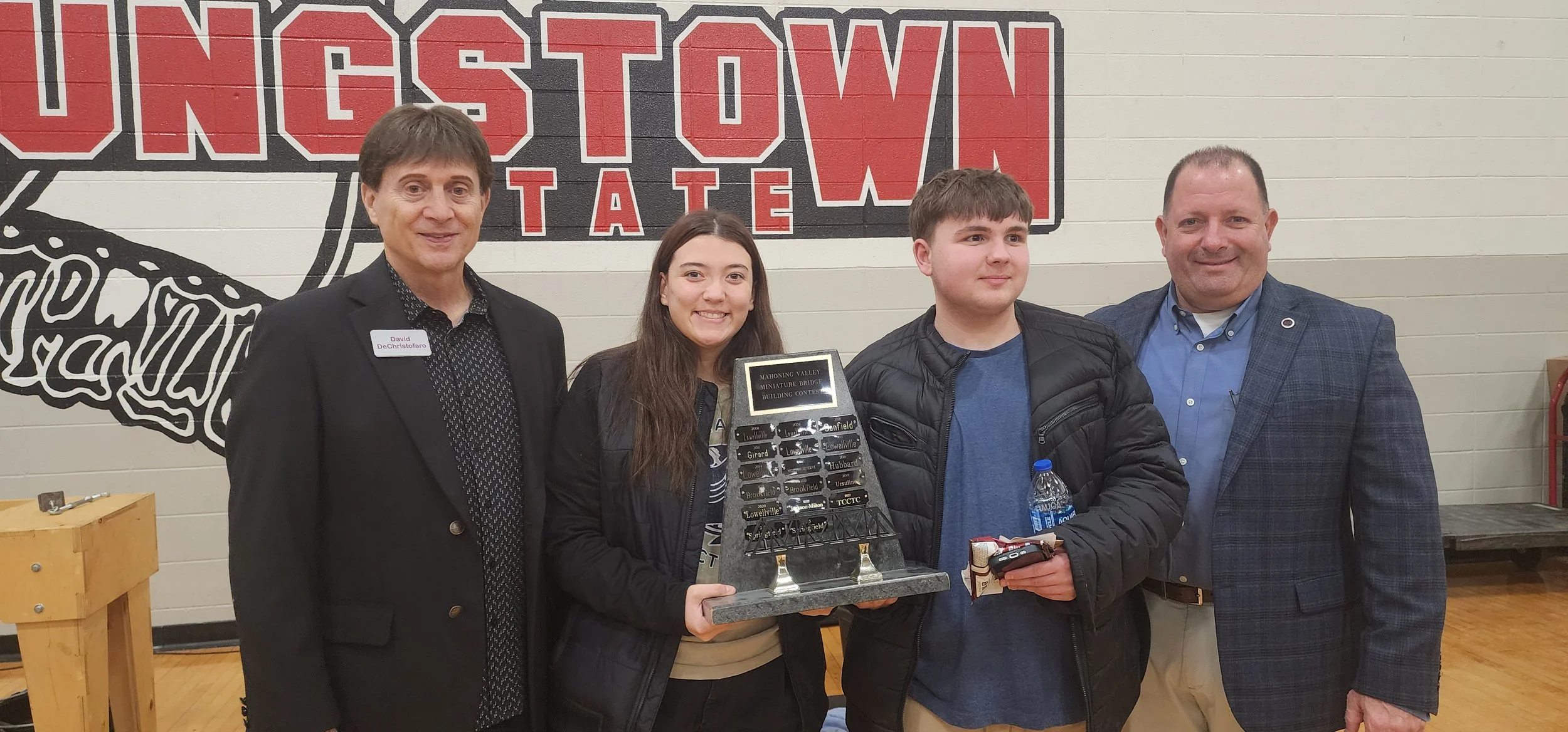 Group of four people, including a teenage girl holding a miniature building contest award, posing in front of a wall with 'Ingestown' sign, with two men and a boy, inside a gymnasium or community center.