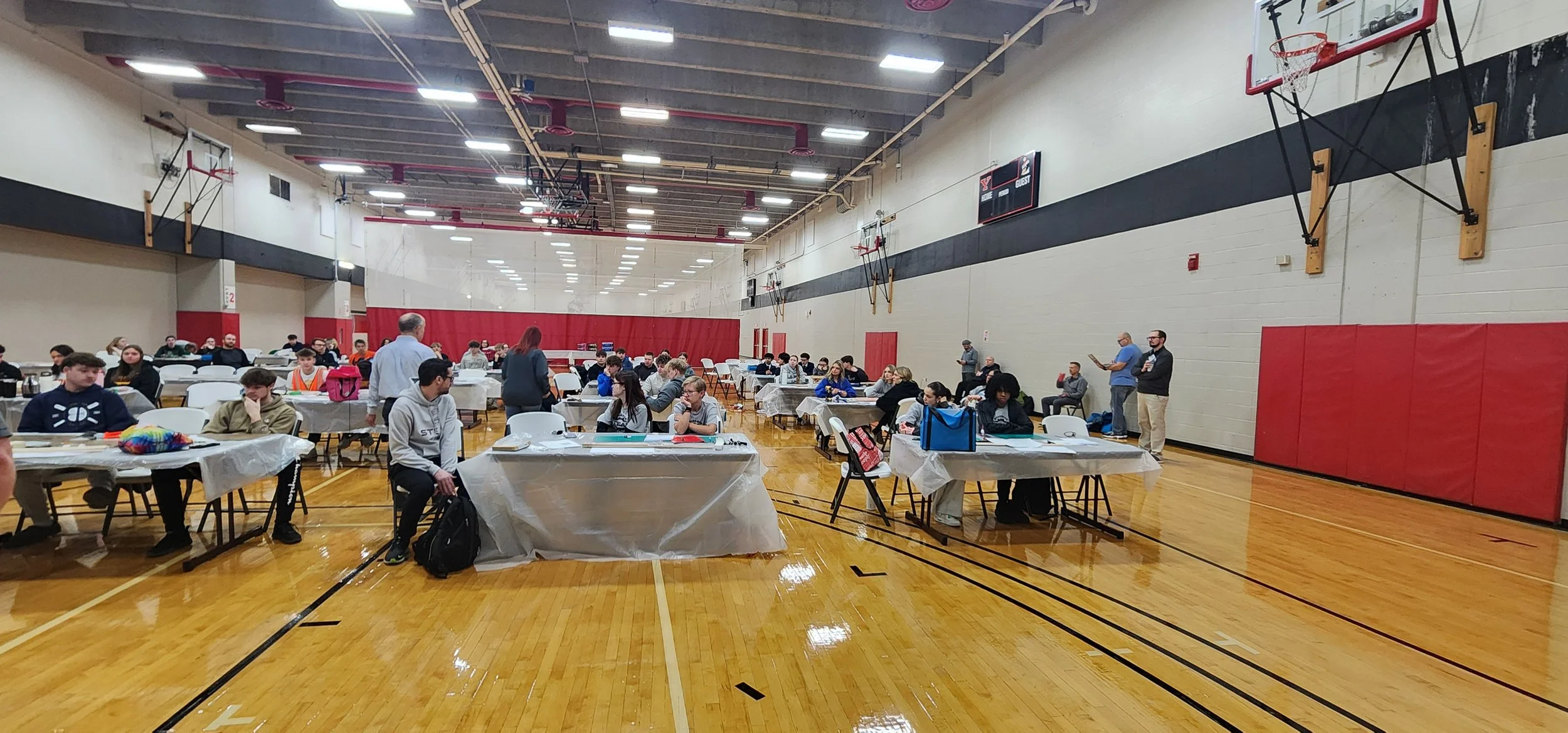 A large gymnasium filled with people sitting at tables covered with white tablecloths, some standing and walking around. The gym has a polished wooden floor, basketball hoops on the walls, and a scoreboard hanging from the ceiling.