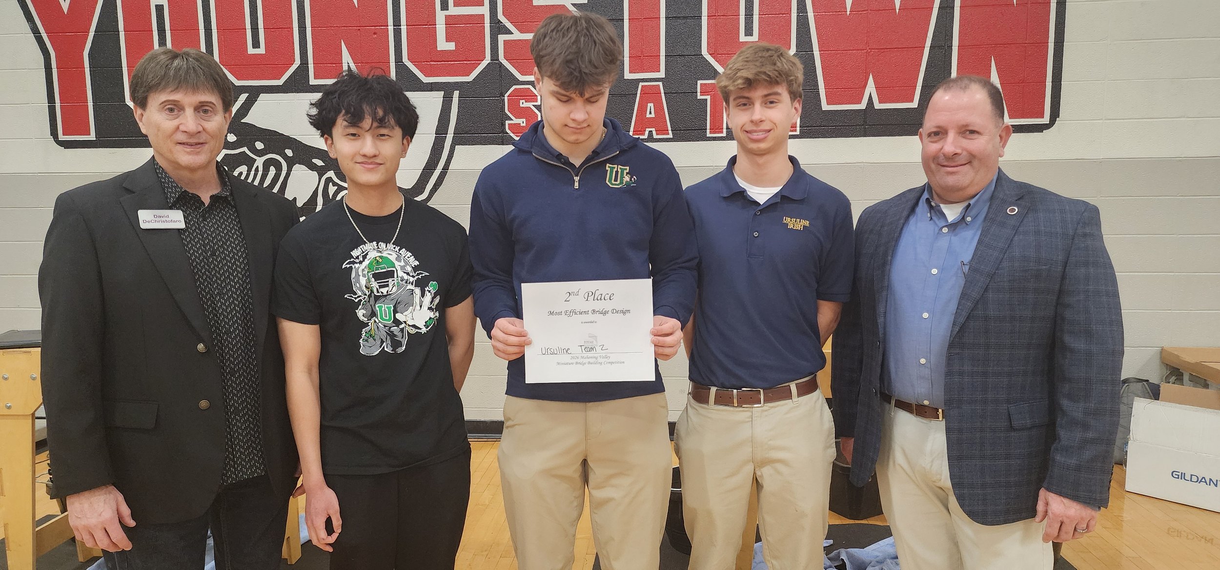 Group of five people, three young men and two older men, standing together indoors in front of a wall with a large red and black graphic. The young man in the middle is holding a certificate for second place in the Most Efficient Bridge Design catego