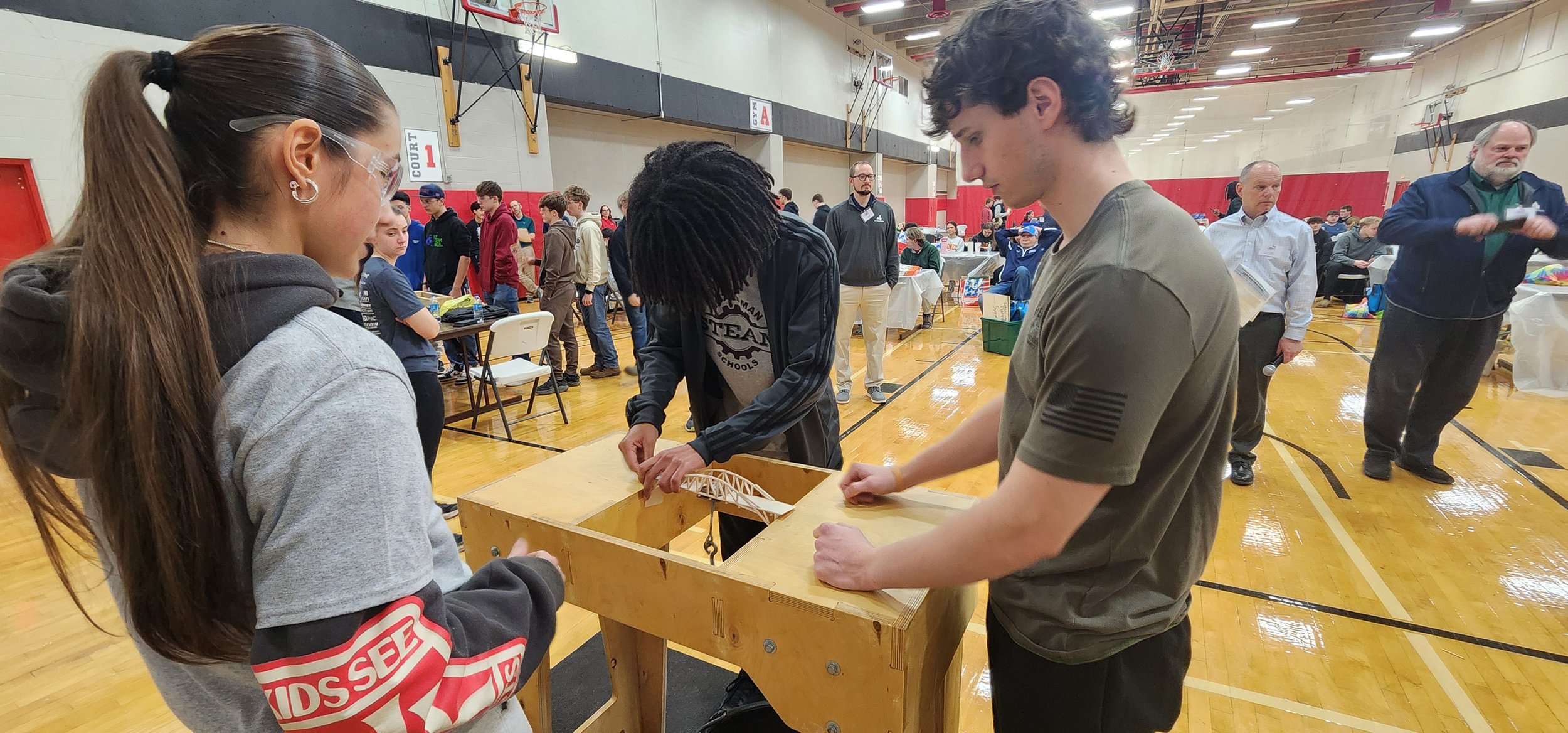 A group of students and adults in a gymnasium participating in a STEM activity, with some working on a bridge project inside a wooden box, and others observing or walking around. There are tables, chairs, and gym equipment visible in the background.