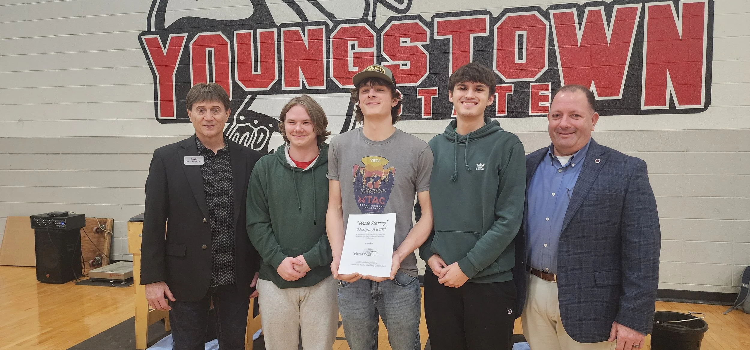 Group of five people standing indoors in front of a wall with a large YMCA Youngstown logo, with three young men in the middle holding a certificate, flanked by two adult men, one on each side.